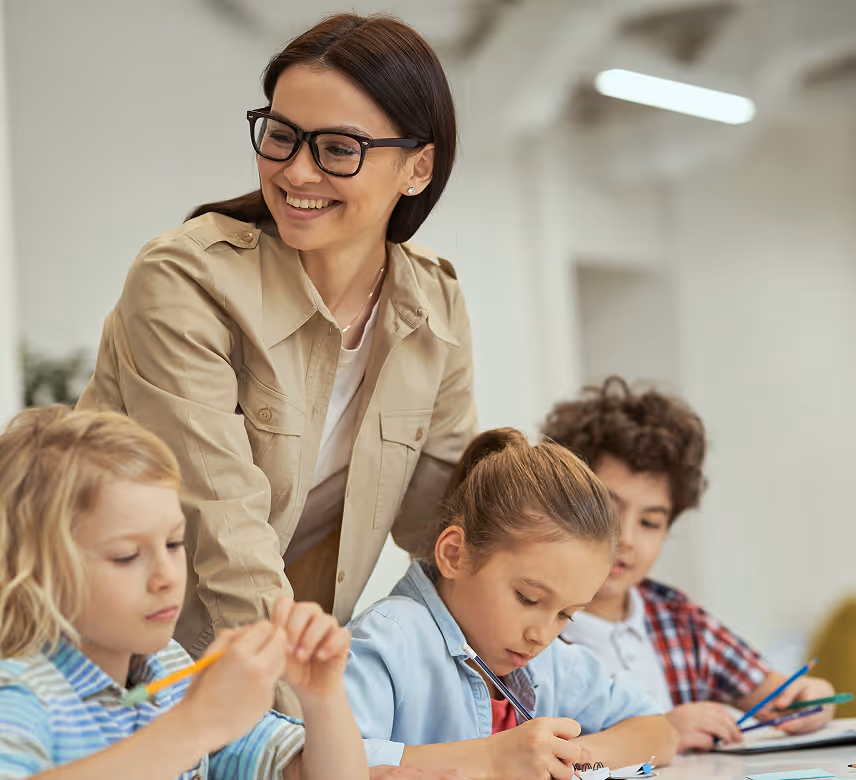 Une enseignante souriante aide trois enfants concentrés sur leur travail à la table.