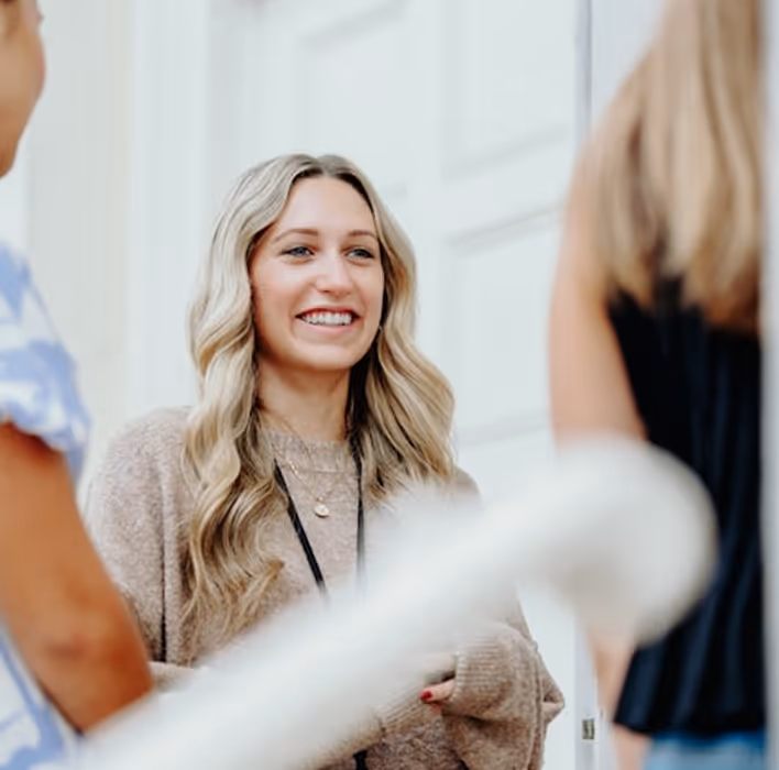 Une femme souriante aux cheveux longs blonds parlant avec deux autres personnes floues.