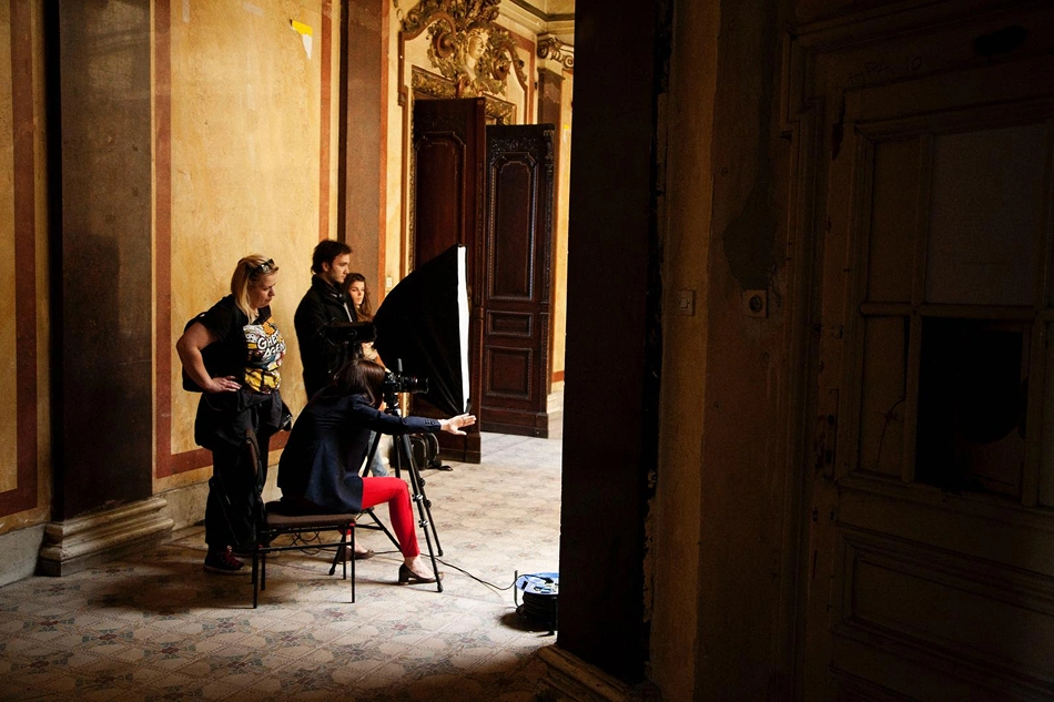 Four people in a historic-looking room with decorated walls; one person is seated operating a camera on a tripod with lighting equipment, and the others are standing nearby observing.