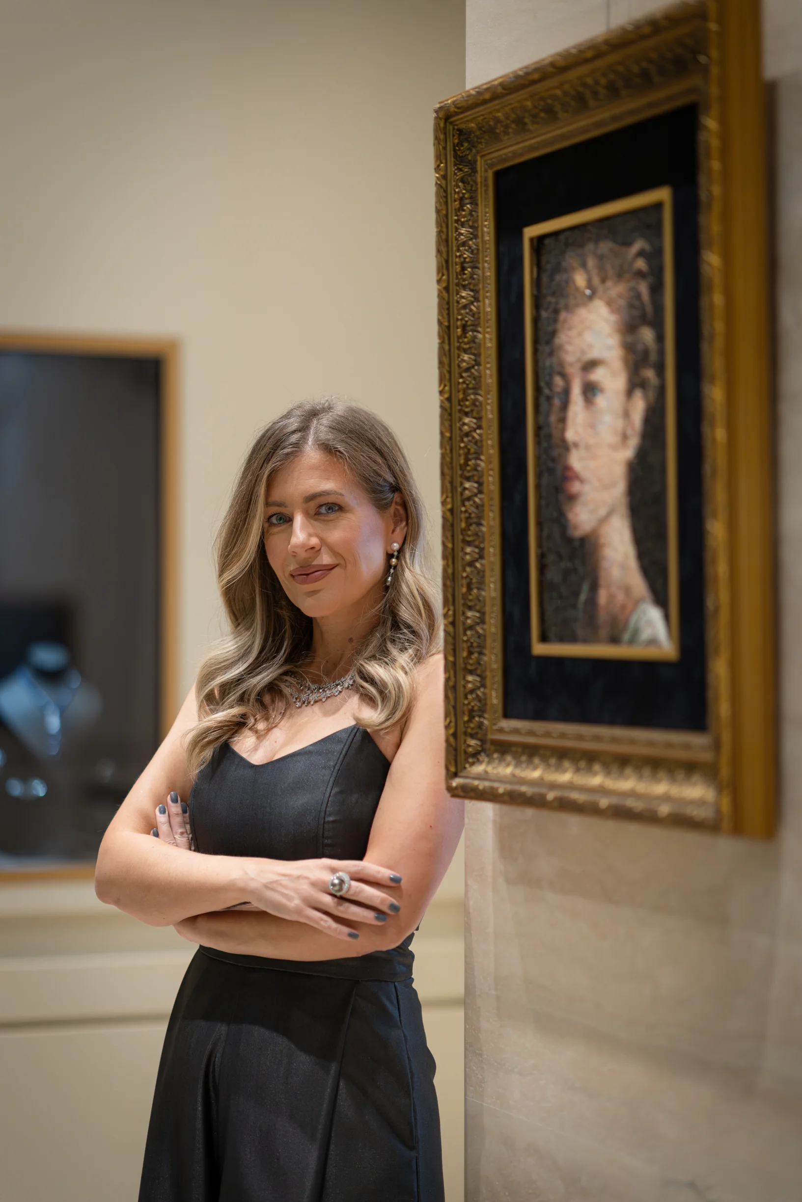 Woman in elegant black dress standing with arms crossed beside a framed portrait in a gallery.