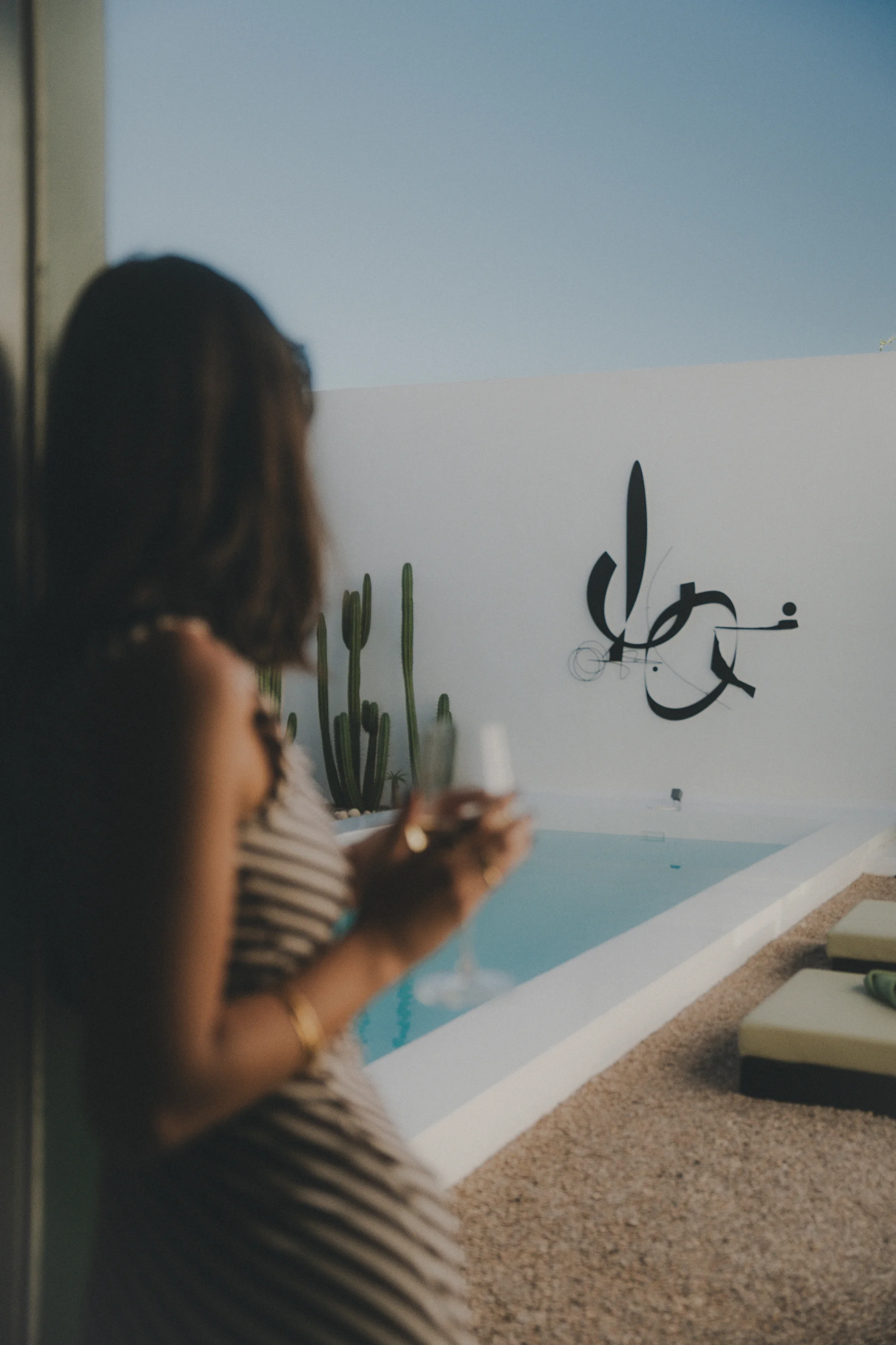Woman holding a glass, overlooking a modern pool with cactus and abstract wall art.