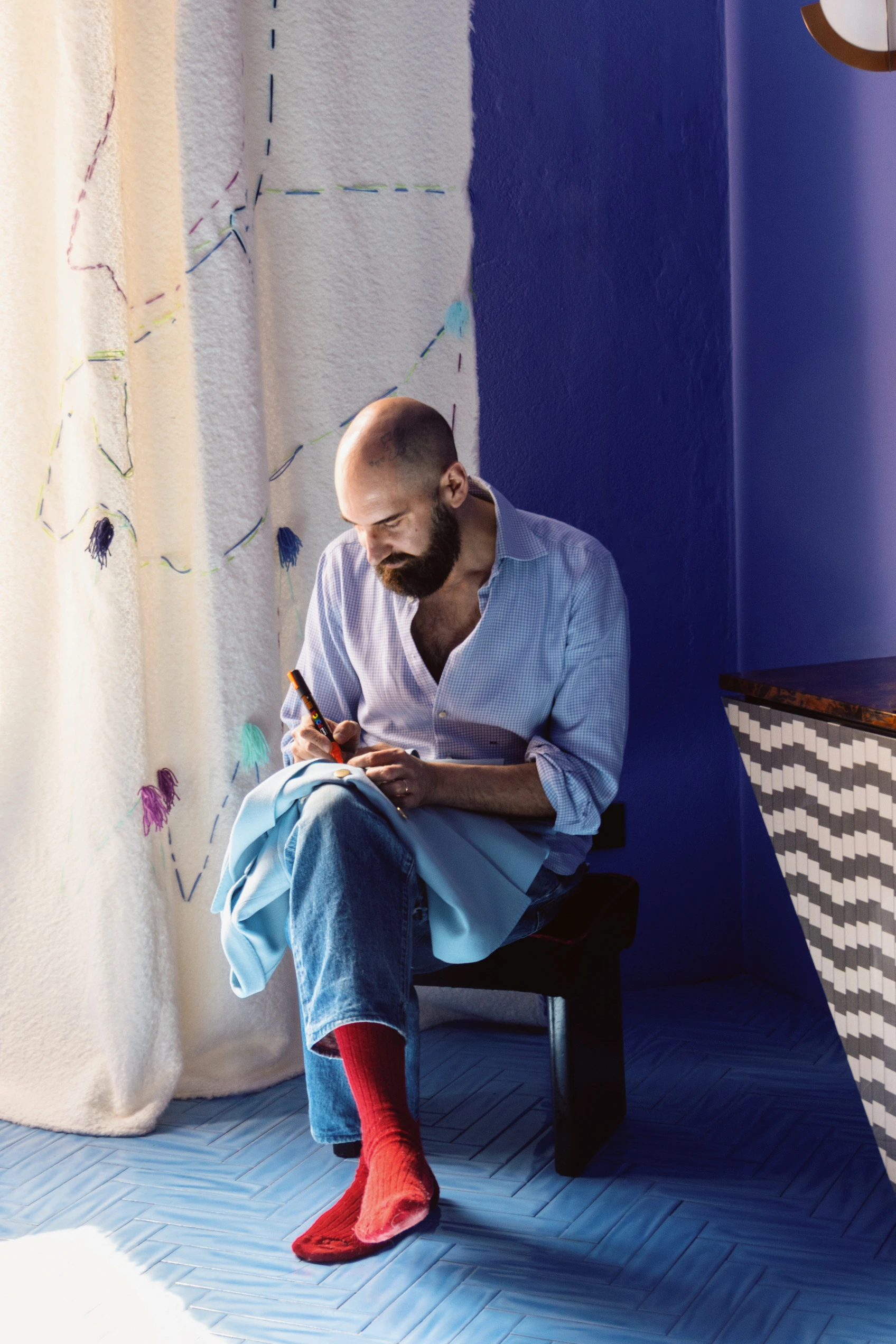 Michele Chiocciolini sitting on a stool, writing on blue fabric by a curtain and blue wall.