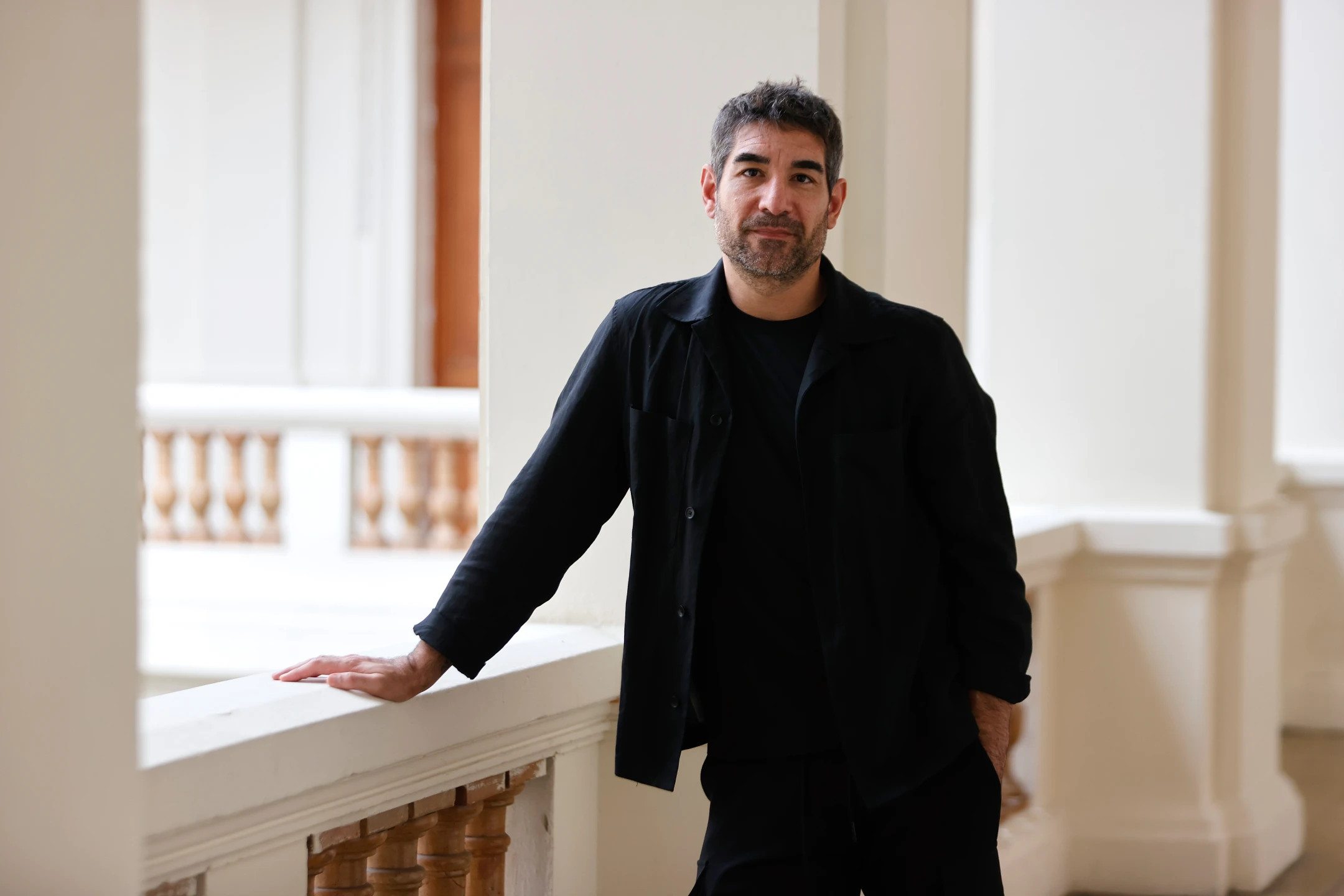 Solimán López in black clothing leaning on a white balcony railing indoors.