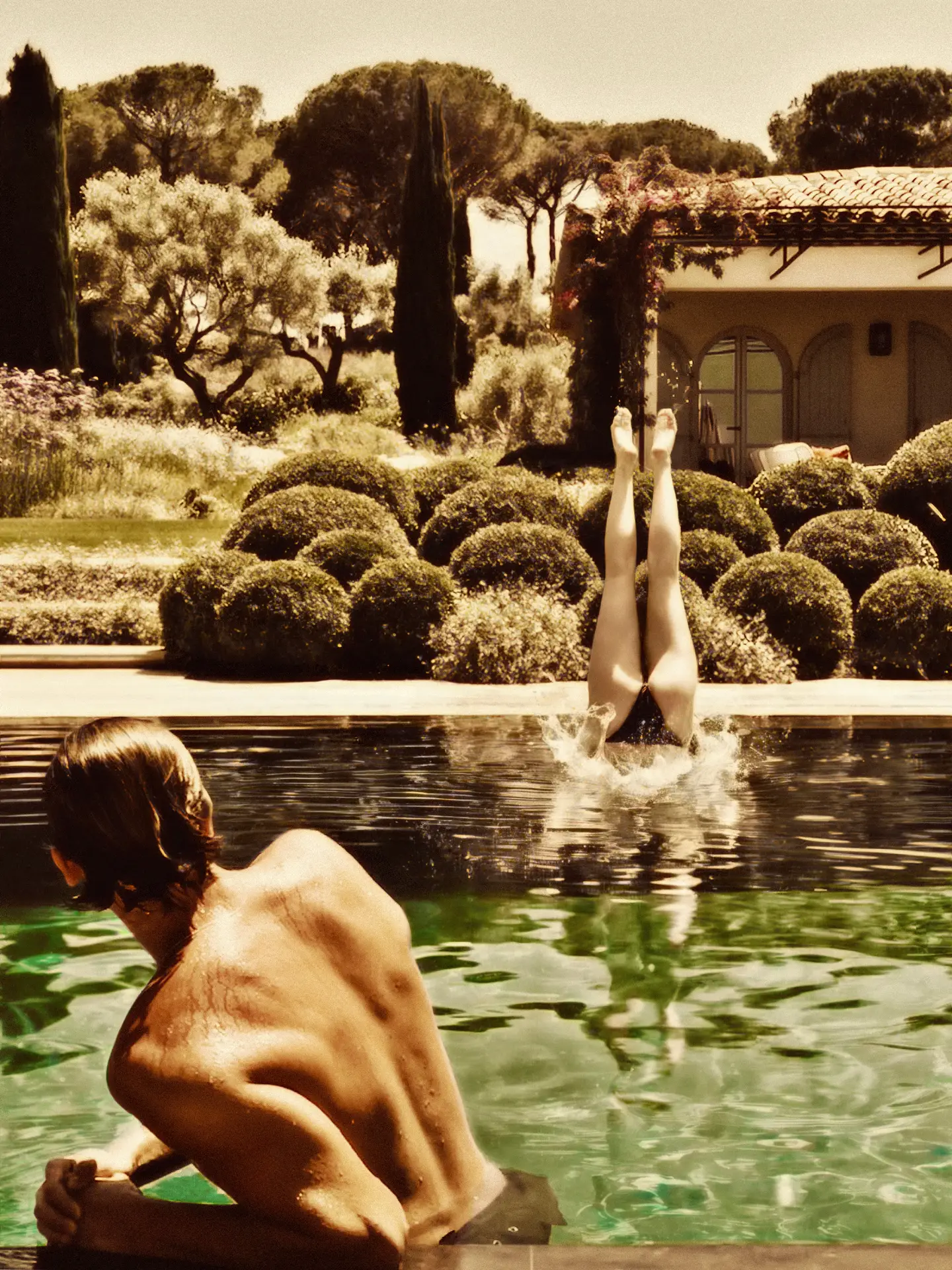 Person with bare back in pool watching another diving headfirst into water with legs up