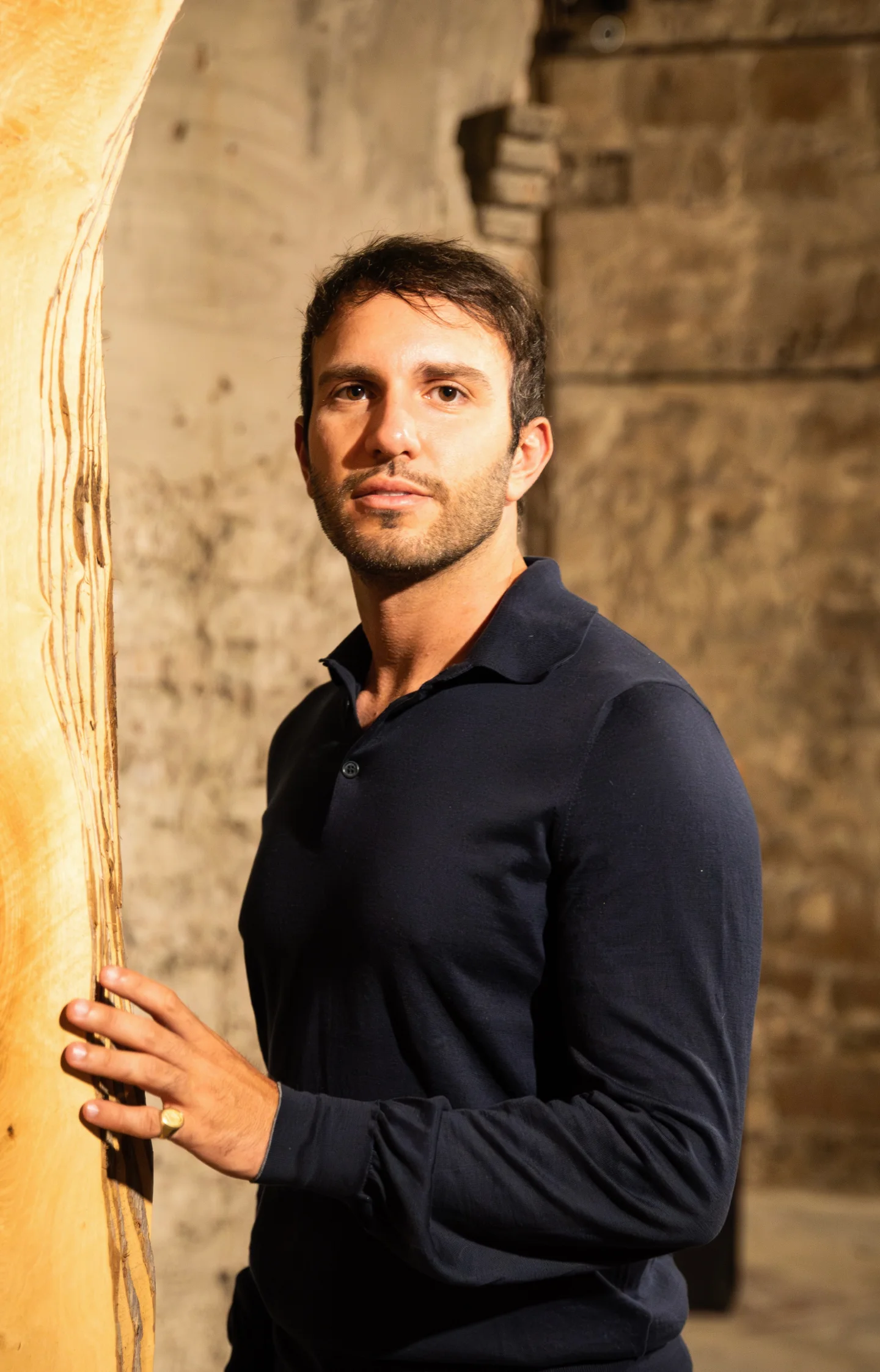 Riccardo Freddo in navy shirt standing indoors, resting hand on light wood panel, stone wall background.