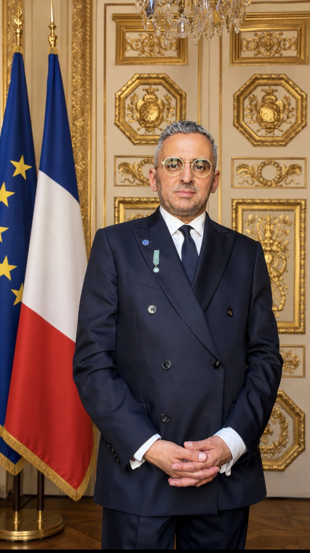 Sid Rouis in a dark suit and glasses stands in front of French and European Union flags with ornate golden decor.
