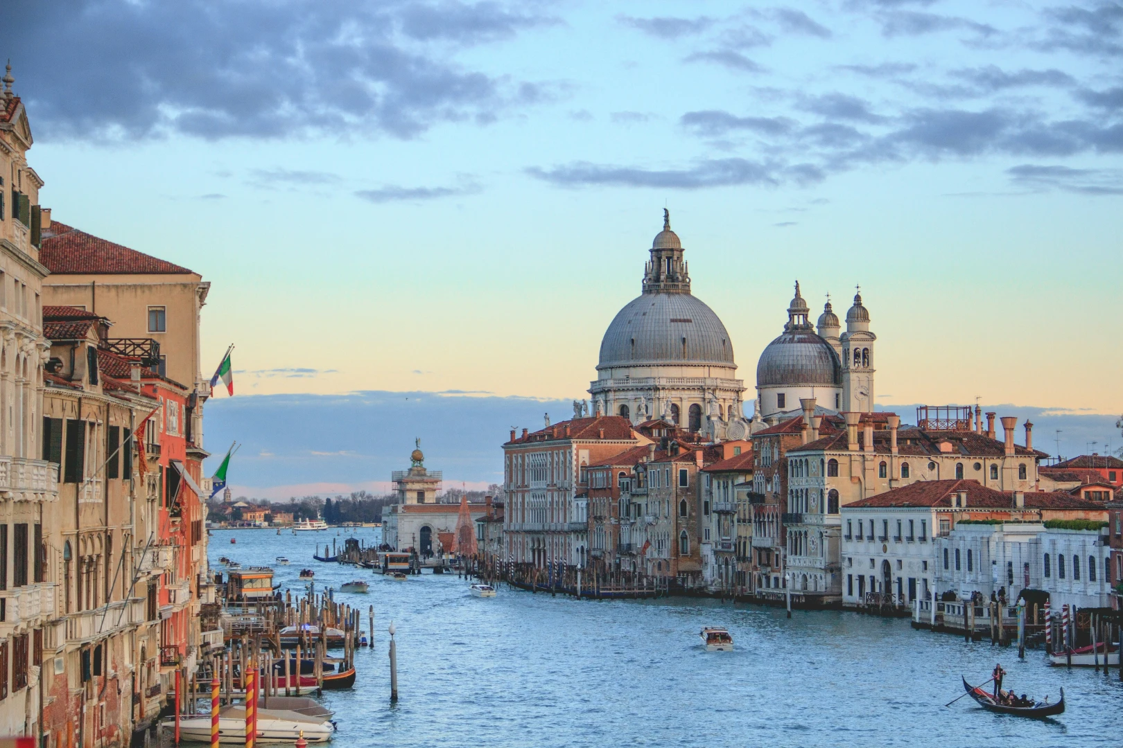 Venice canal with boats and gondola passing historic buildings and domed church at sunset.