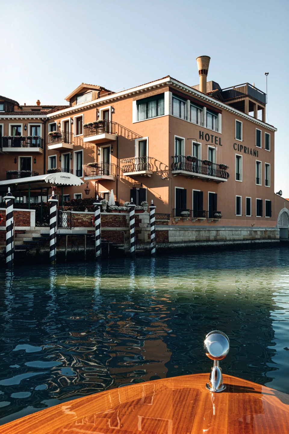 View of the Hotel Cipriani building in Venice with balconies, flower pots, and striped mooring poles along a canal, seen from a wooden boat.