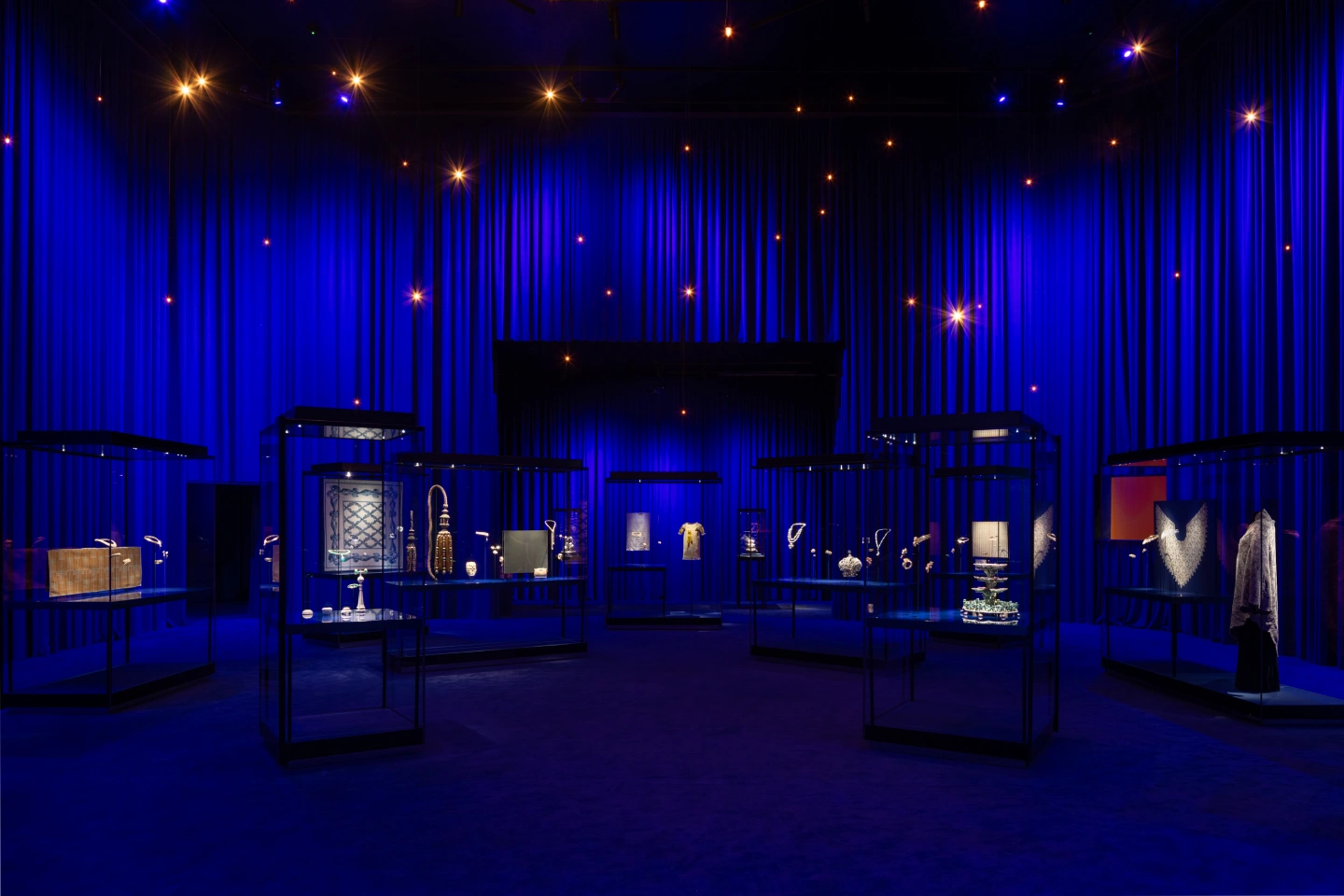 Dimly lit exhibition room with deep blue curtains and floor, featuring multiple glass display cases illuminated to showcase various artifacts and jewelry pieces, with small star-like lights hanging from the ceiling.
