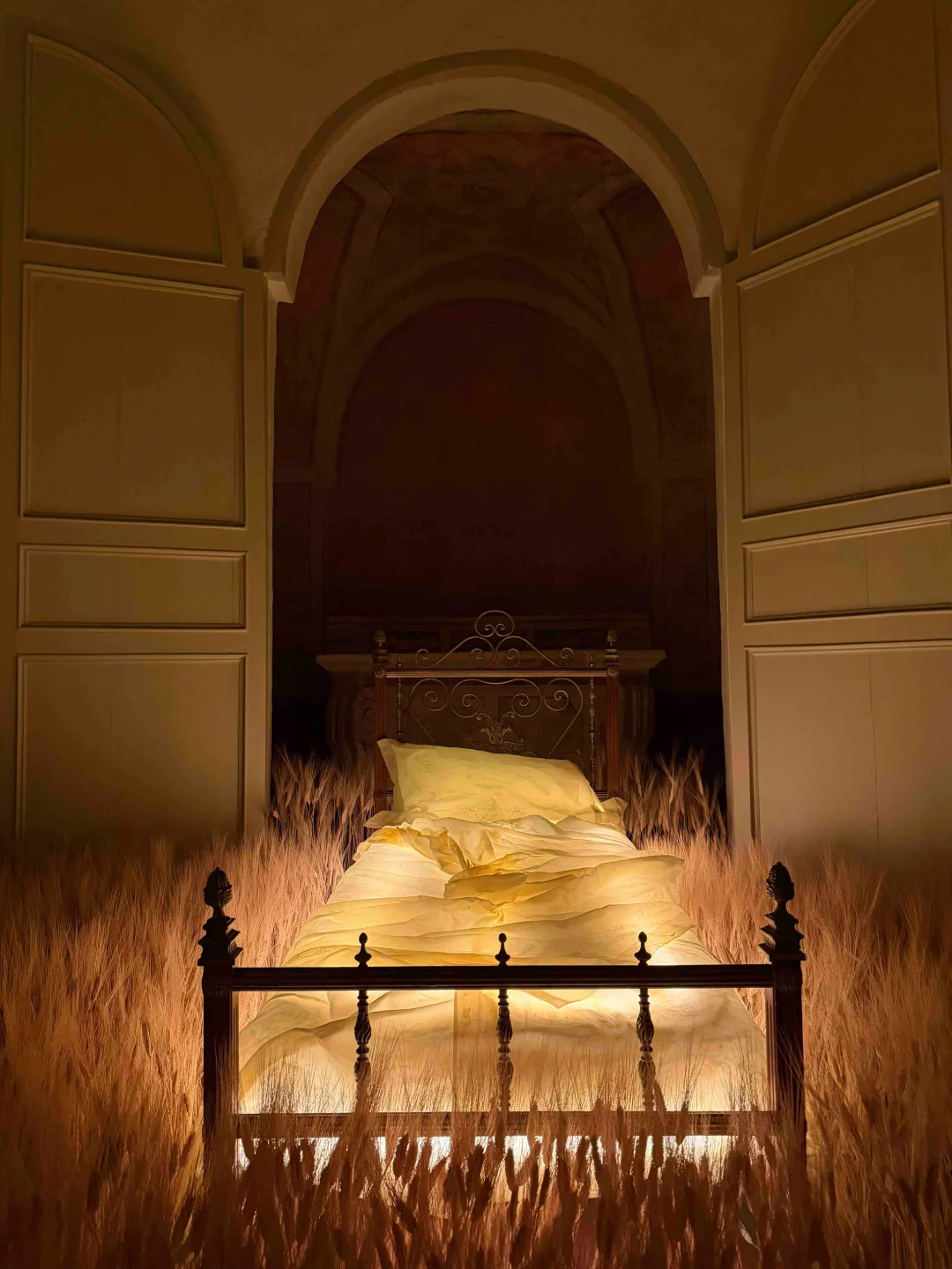 An ornate iron bed with illuminated yellow bedding placed in the middle of a field of tall wheat stalks inside a dimly lit room with arched ceiling and large open wooden doors.