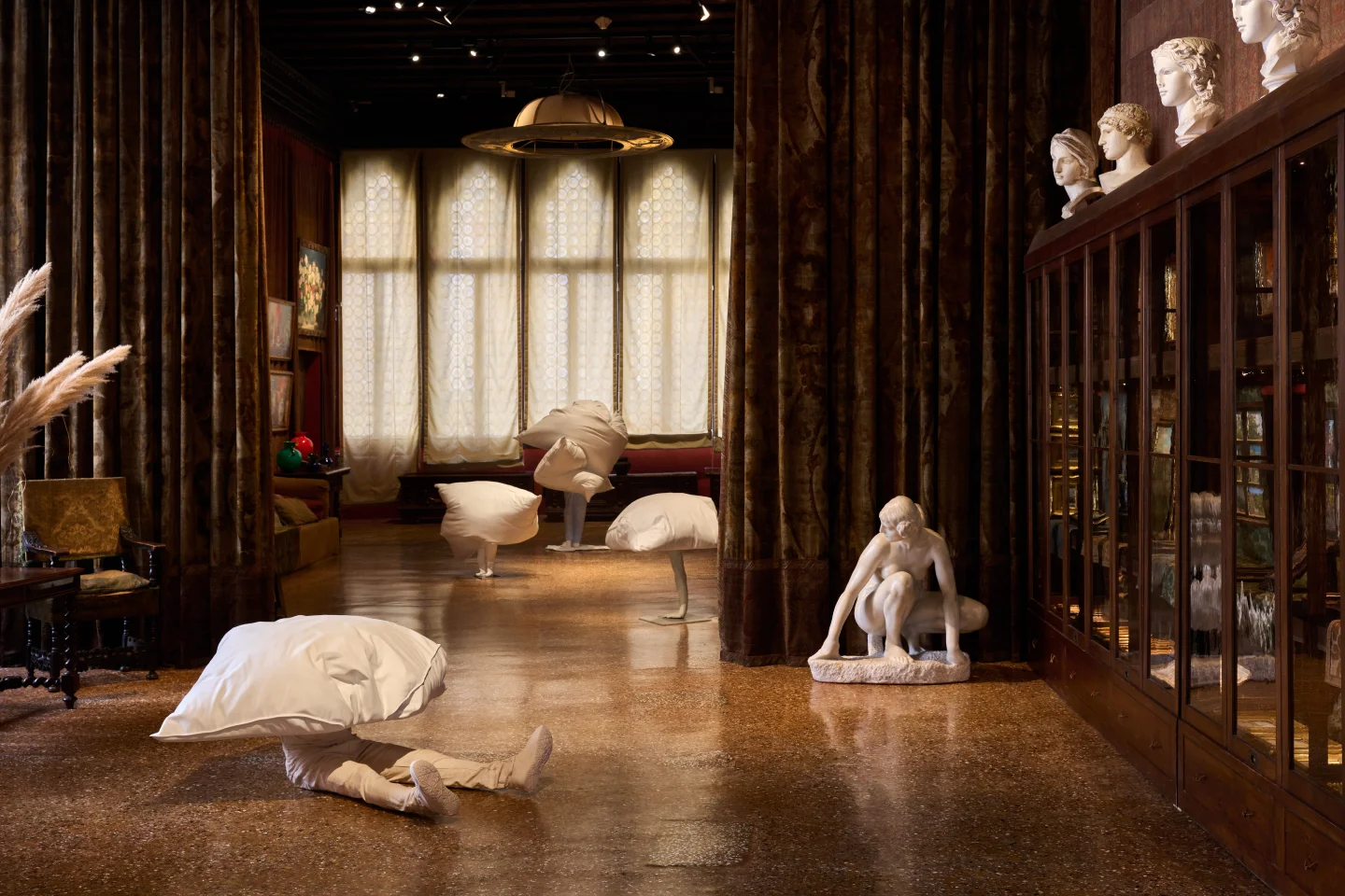 Erwin Wurm art installation in a dimly lit room with dark curtains, large windows with patterned shades, and wooden furniture; several figures covered with large white pillow-like coverings concealing their upper bodies are positioned on the floor and standing; a white classical-style sculpture of a crouching figure is near a glass-fronted wooden cabinet topped with classical busts.