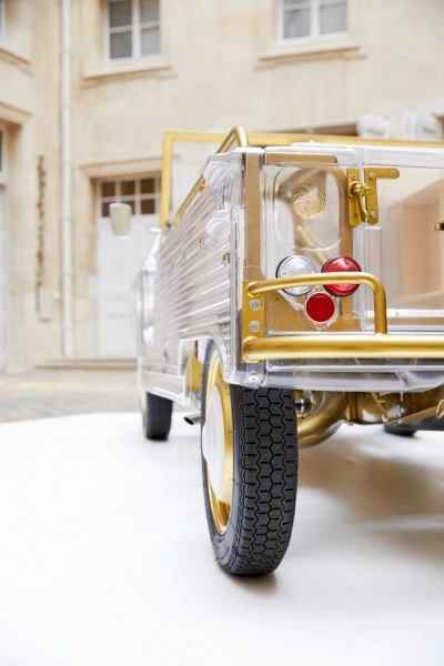 Rear-left view of a transparent model vehicle with gold and white details, showing wheels, tail lights, and inner mechanical parts, displayed indoors against a stone building background.