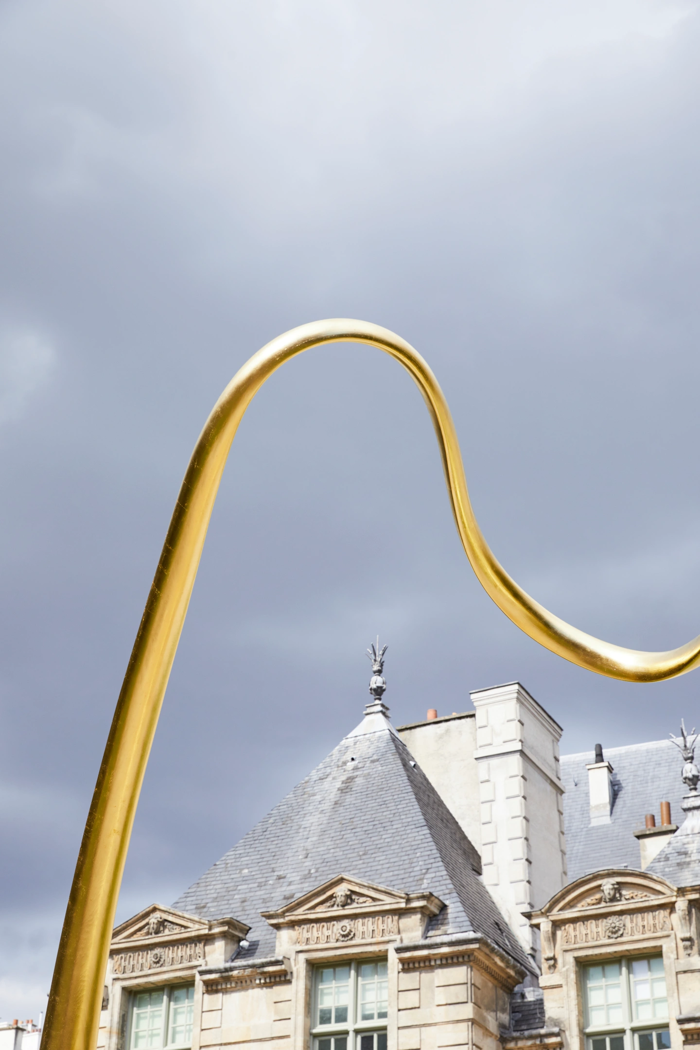 A curved shiny gold sculpture arching over the rooftop of a historic stone building with grey slate roofs and ornate window frames under a cloudy sky.