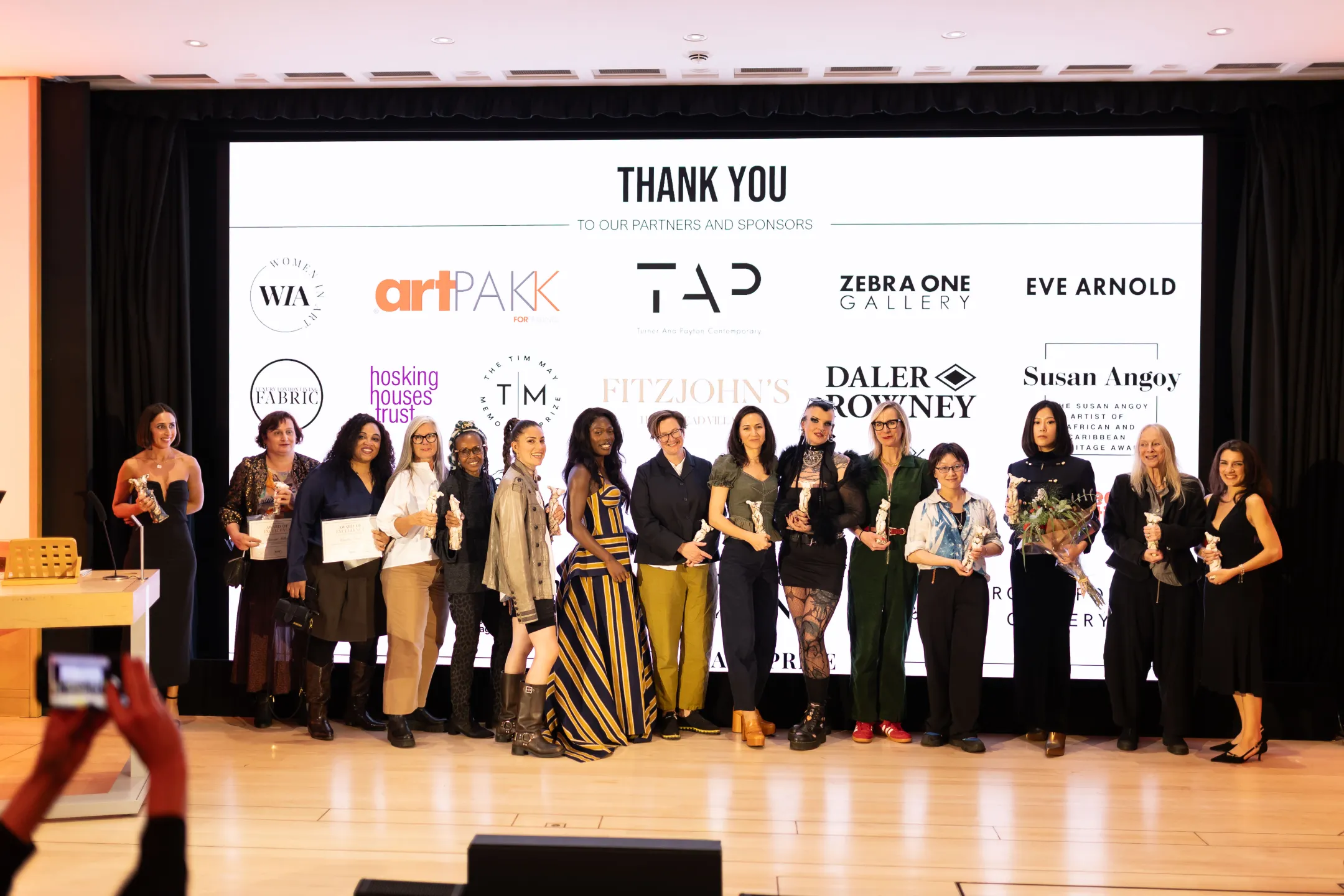 A group of diverse women standing on a stage holding awards and certificates in front of a large screen displaying logos of sponsors and partners, with one person in the foreground taking a photo with a smartphone.