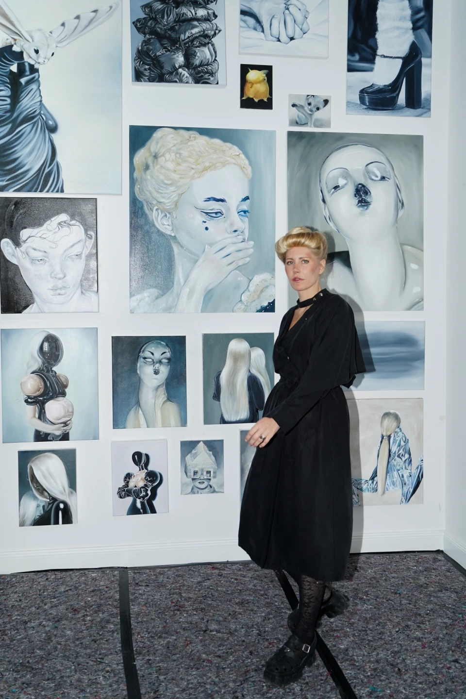 Woman with blond hair wearing a black dress and patterned tights standing in front of a white wall displaying a collection of surreal and abstract paintings featuring human faces, figures, and objects in muted blue and grey tones.