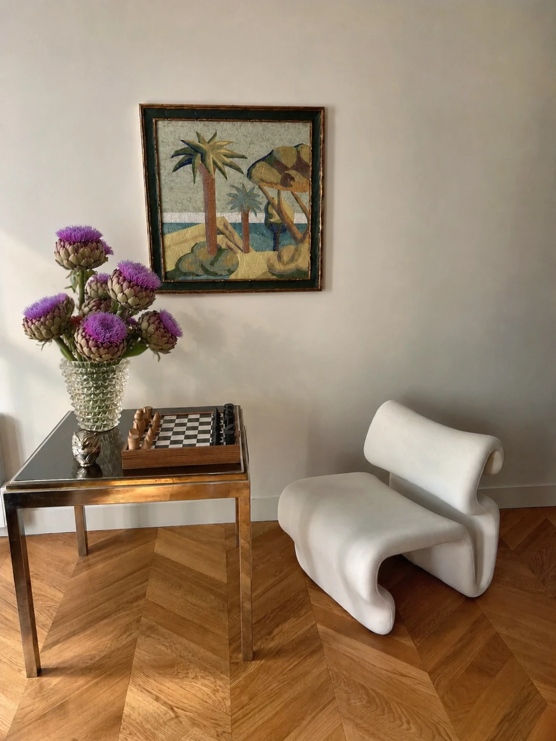 Minimalist interior with a modern white curved chair on herringbone wooden floor beside a reflective square table holding a glass vase with purple thistle flowers and a wooden chess set; above, a framed artwork depicting stylized palm trees and sand.