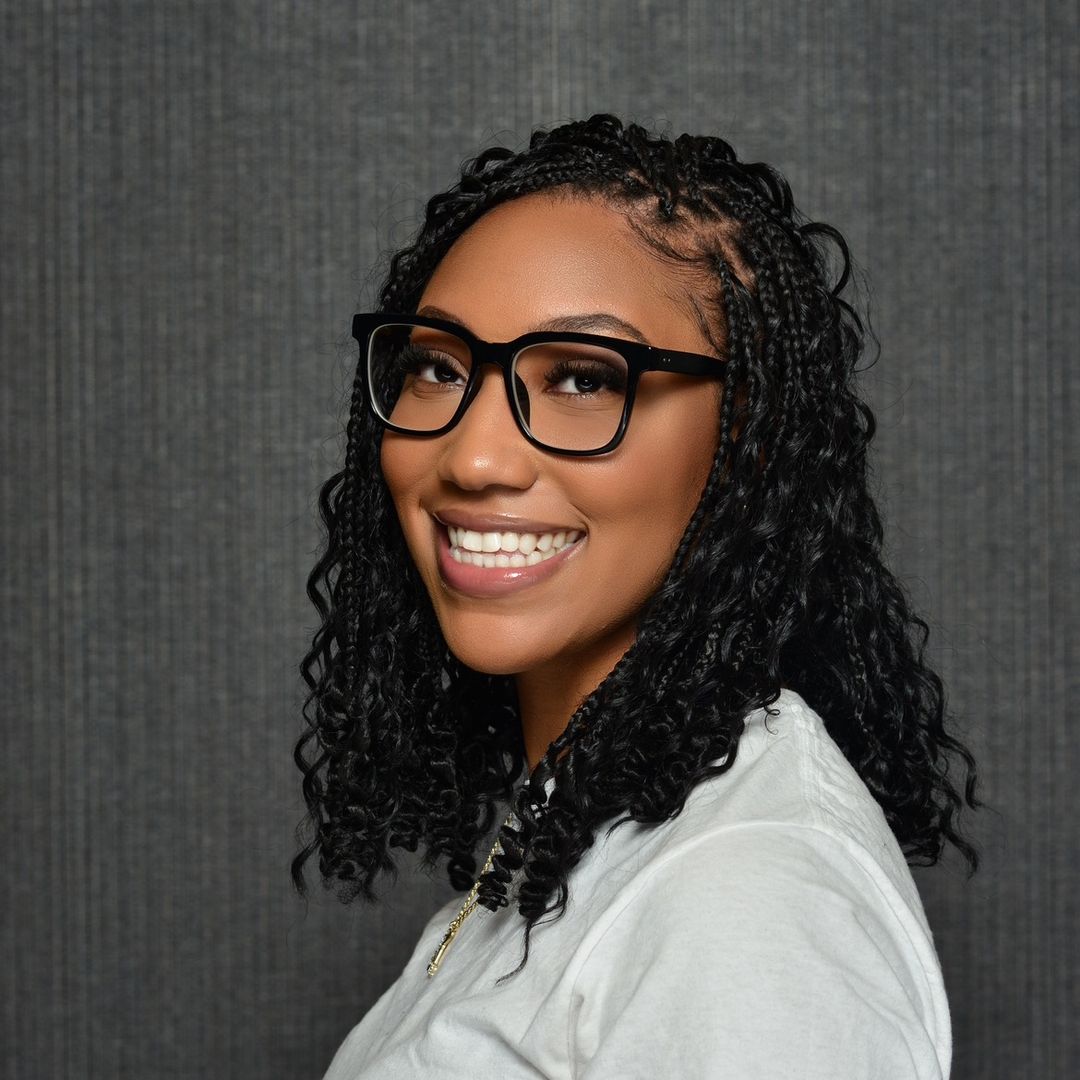 Smiling woman with black braided hair and large black glasses wearing a white shirt against a gray textured background.