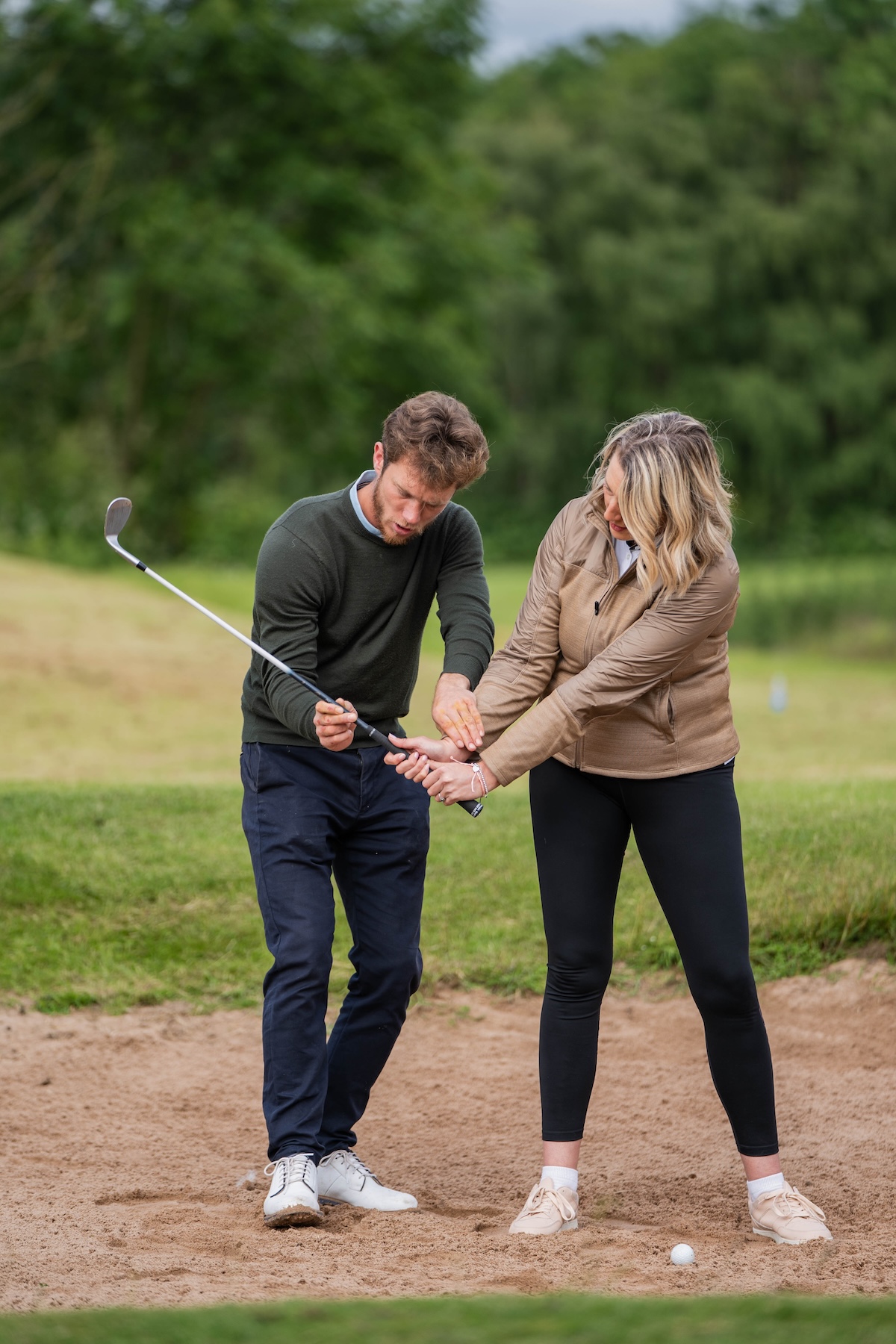 Golf instructor teaching woman how to hold a golf club on a sand bunker with ball in front.