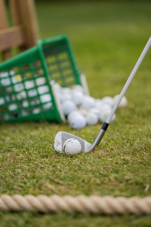 Golf club poised to hit a golf ball on grass with a green basket holding more balls nearby.