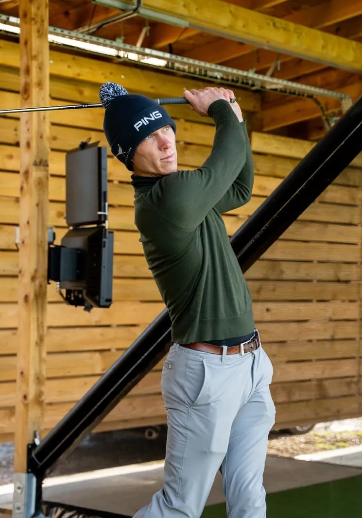 Man in a green sweater and PING hat swinging a golf club indoors at a wooden driving range.