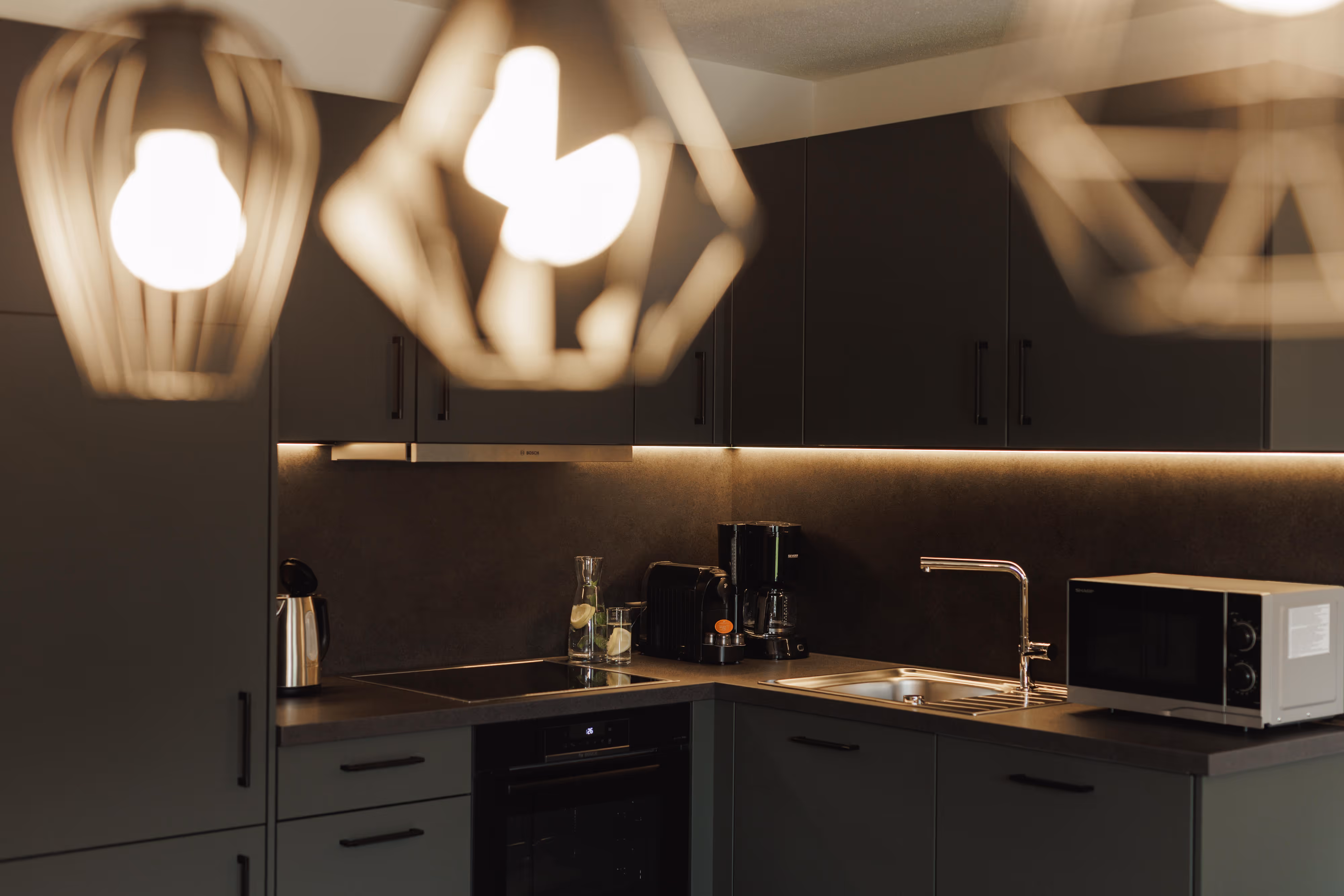 Moderne dunkle Küche mit Küchenutensilien, Kaffeemaschinen, Spüle und Mikrowelle unter warmem Licht.