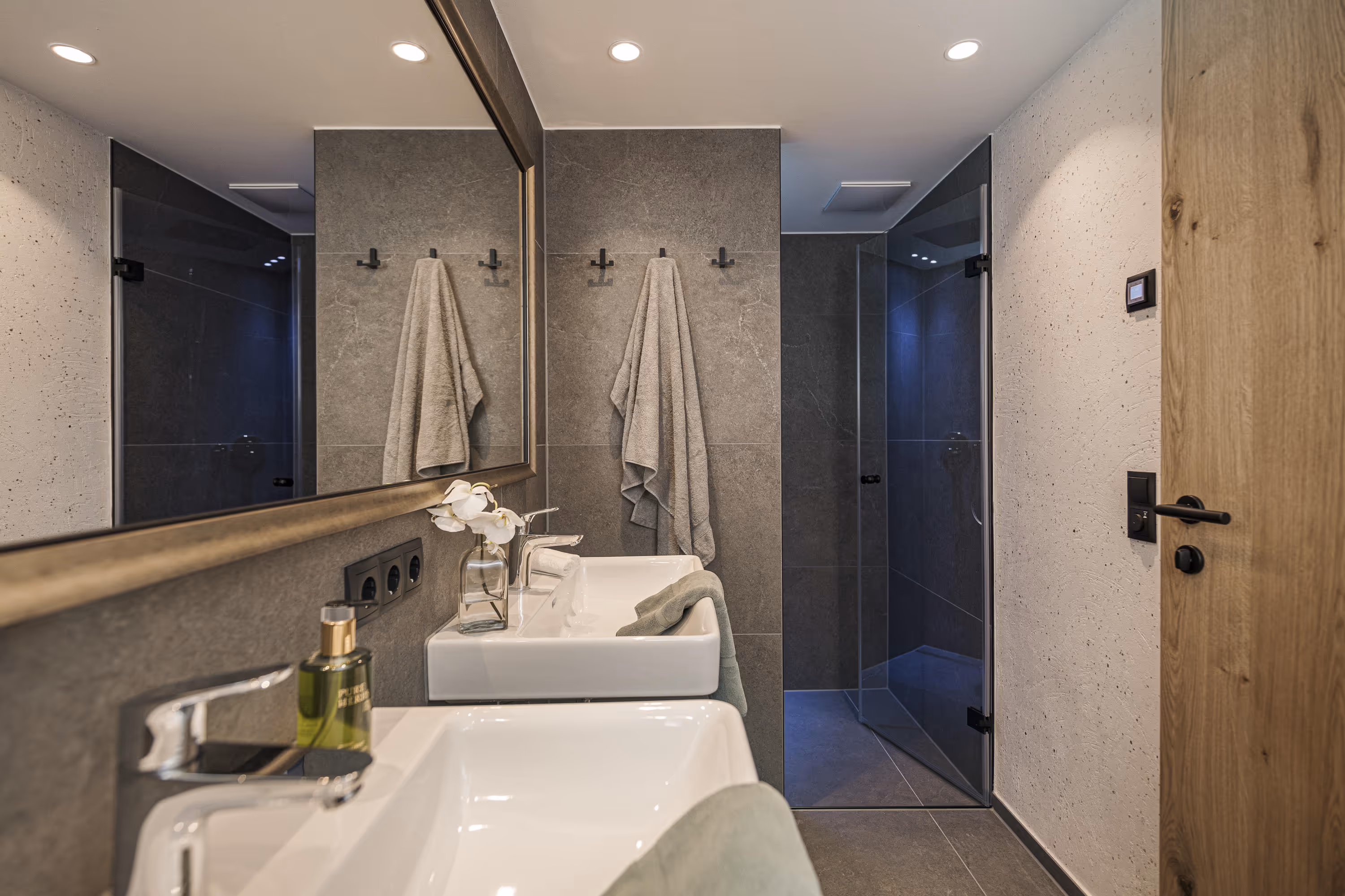 Modern bathroom with double white sinks, large mirror, gray tiles, and a glass door shower.