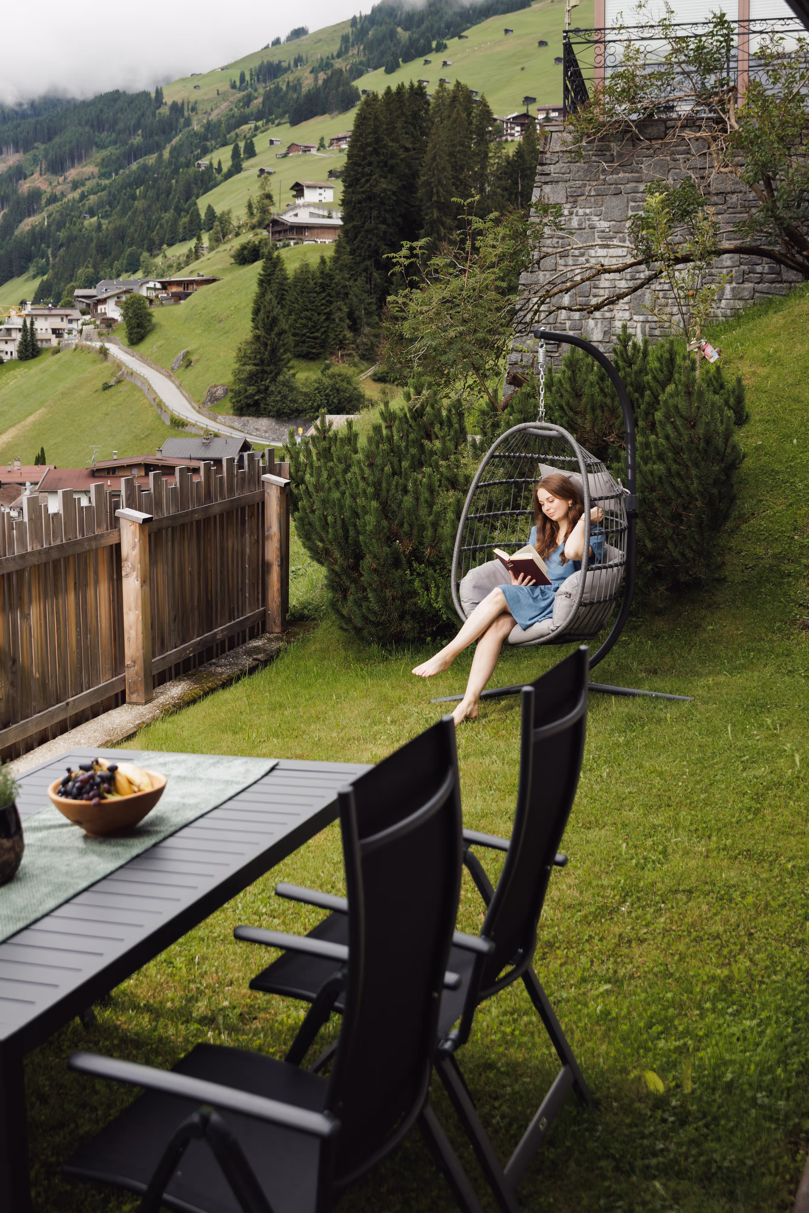 Frau liest auf einem hängenden Korbsessel im Garten mit Berg- und Dorfblick im Hintergrund.