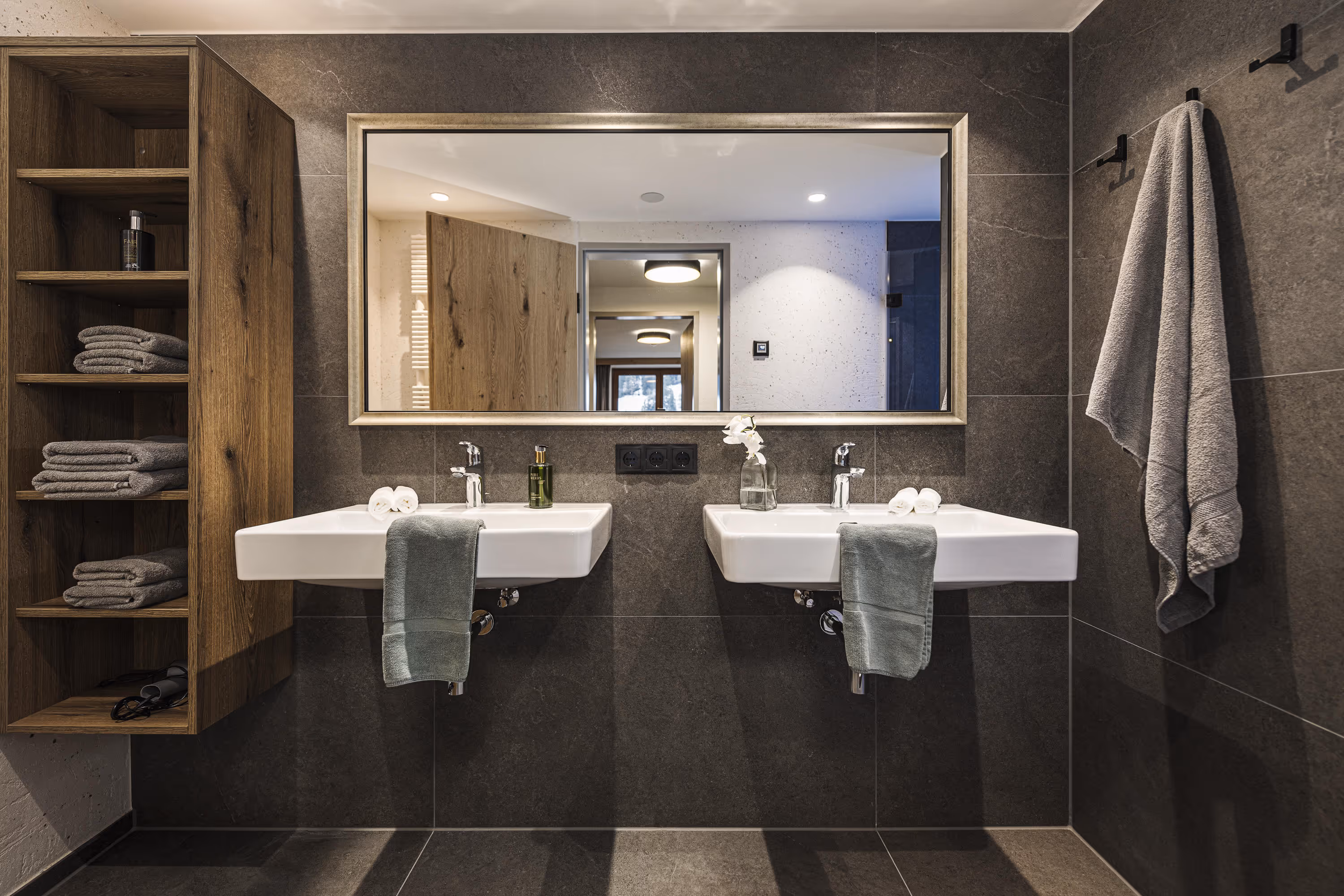 Modern bathroom with two white sinks, large mirror, wooden shelf with towels, and towel hooks on wall.
