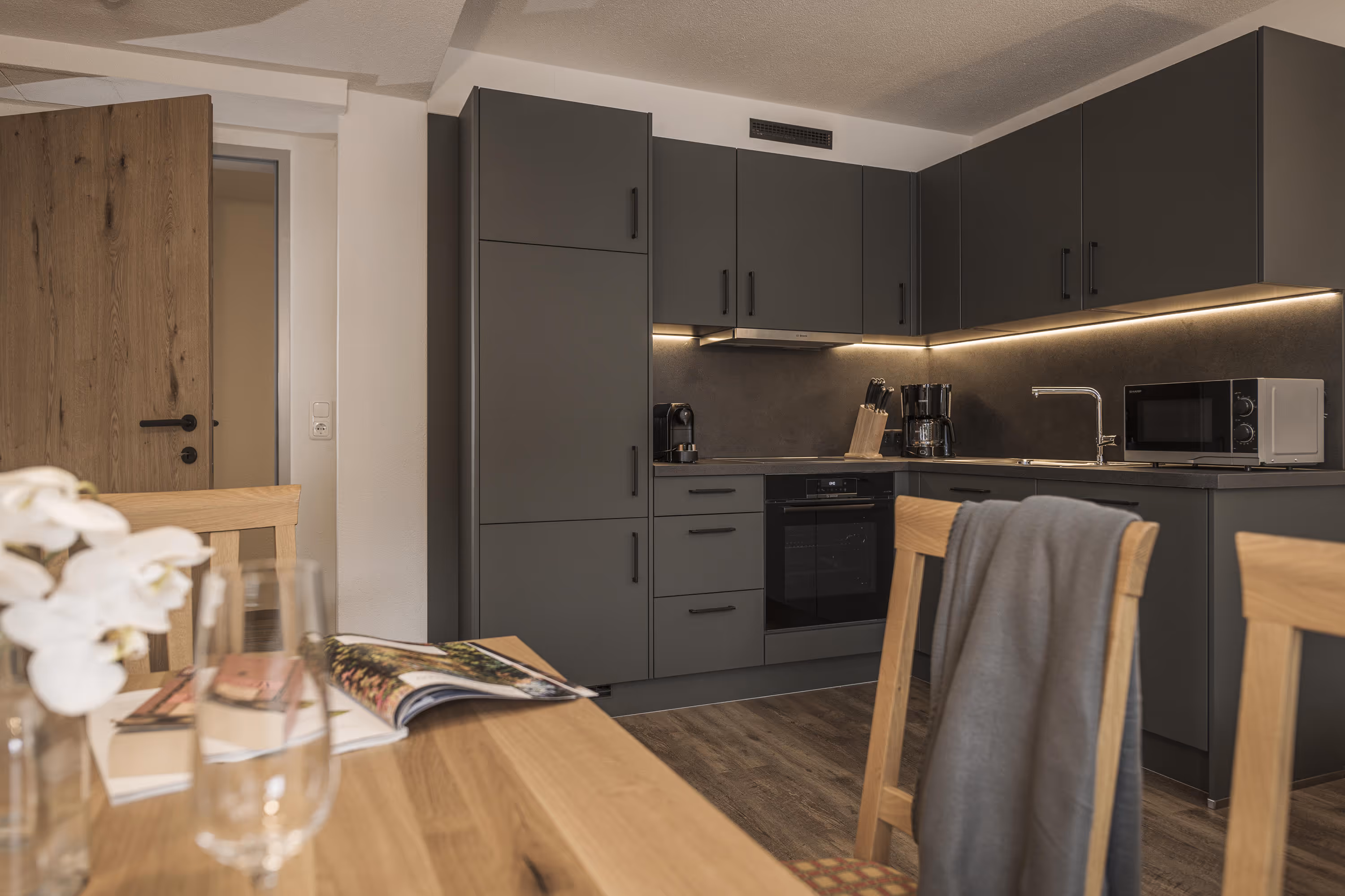 Moderne Küche mit grauen Schränken, Herd, Kaffeemaschine, Mikrowelle und Holztisch mit Stühlen.