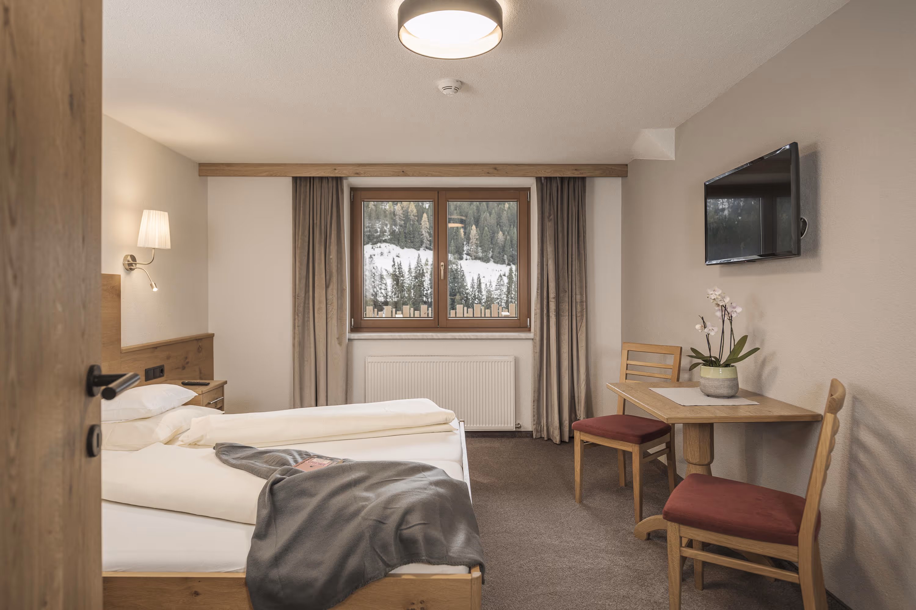 Gemütliches Hotelzimmer mit Bett, Fenster mit Bergblick, Tisch mit Blume und Stühlen, Fernseher an der Wand.