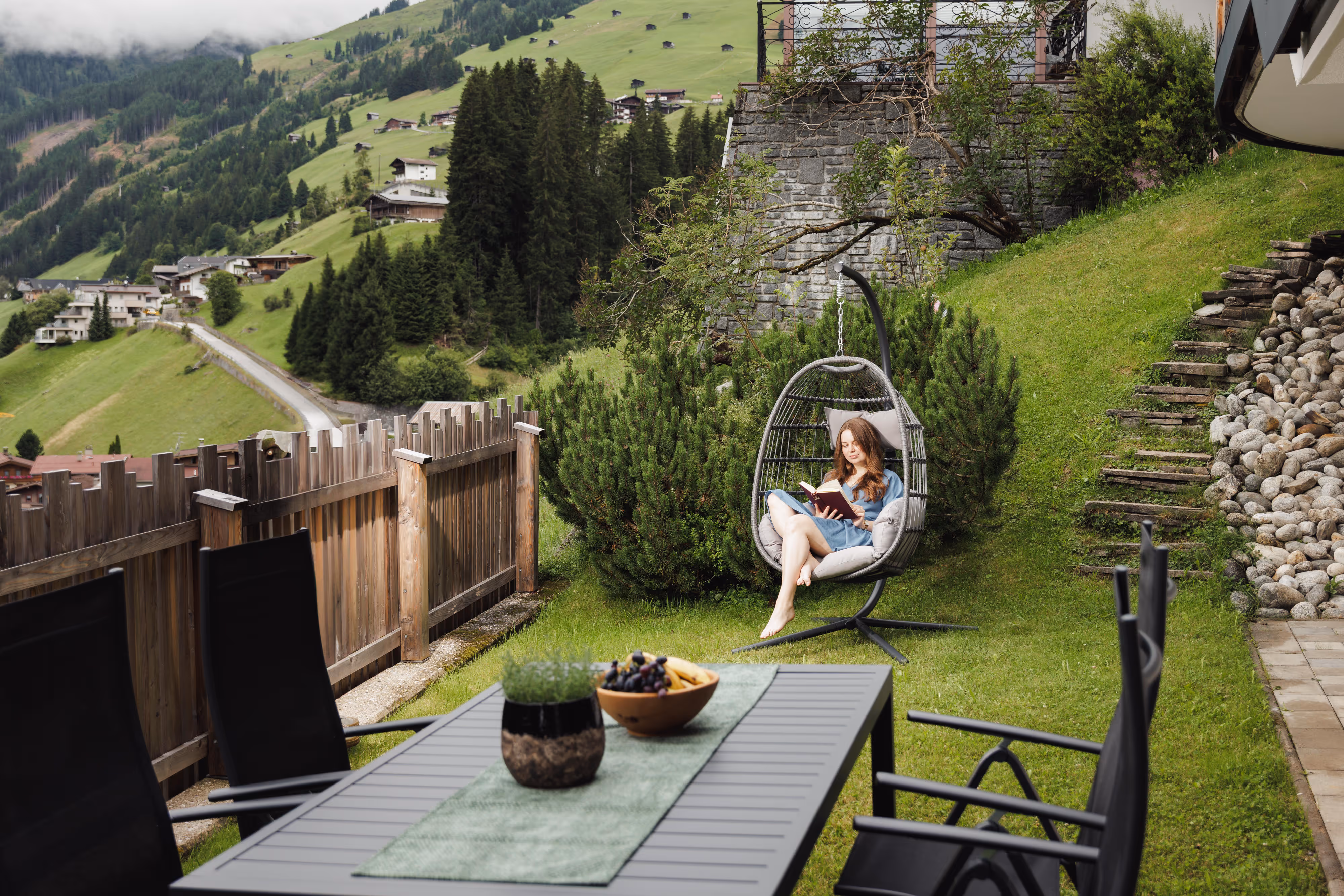 Frau liest entspannt in einem Hängesessel im Garten mit Bergblick und Sitzbereich mit Tisch.