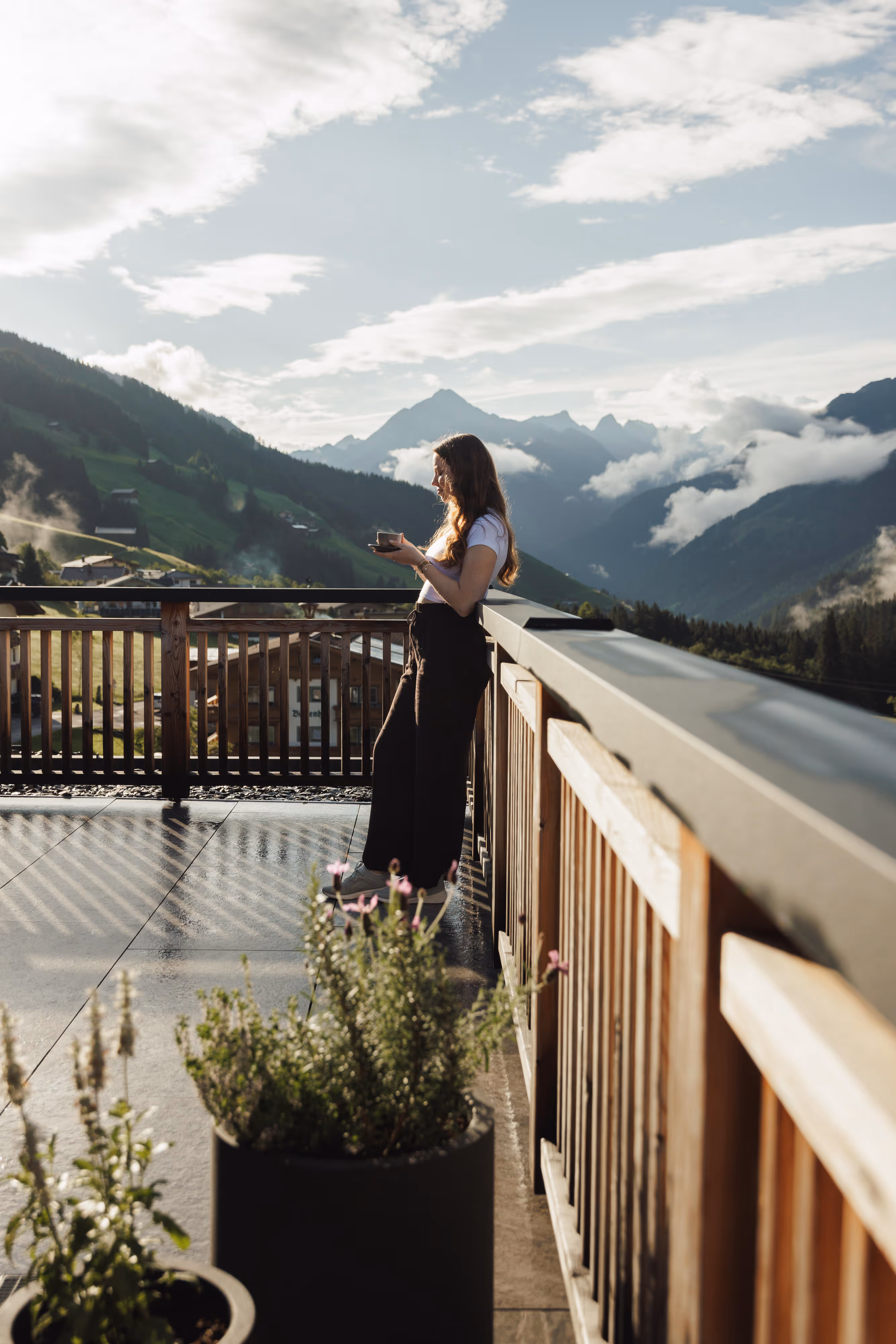 Frau steht mit einer Tasse auf einem Balkon mit Bergblick an einem sonnigen Tag.