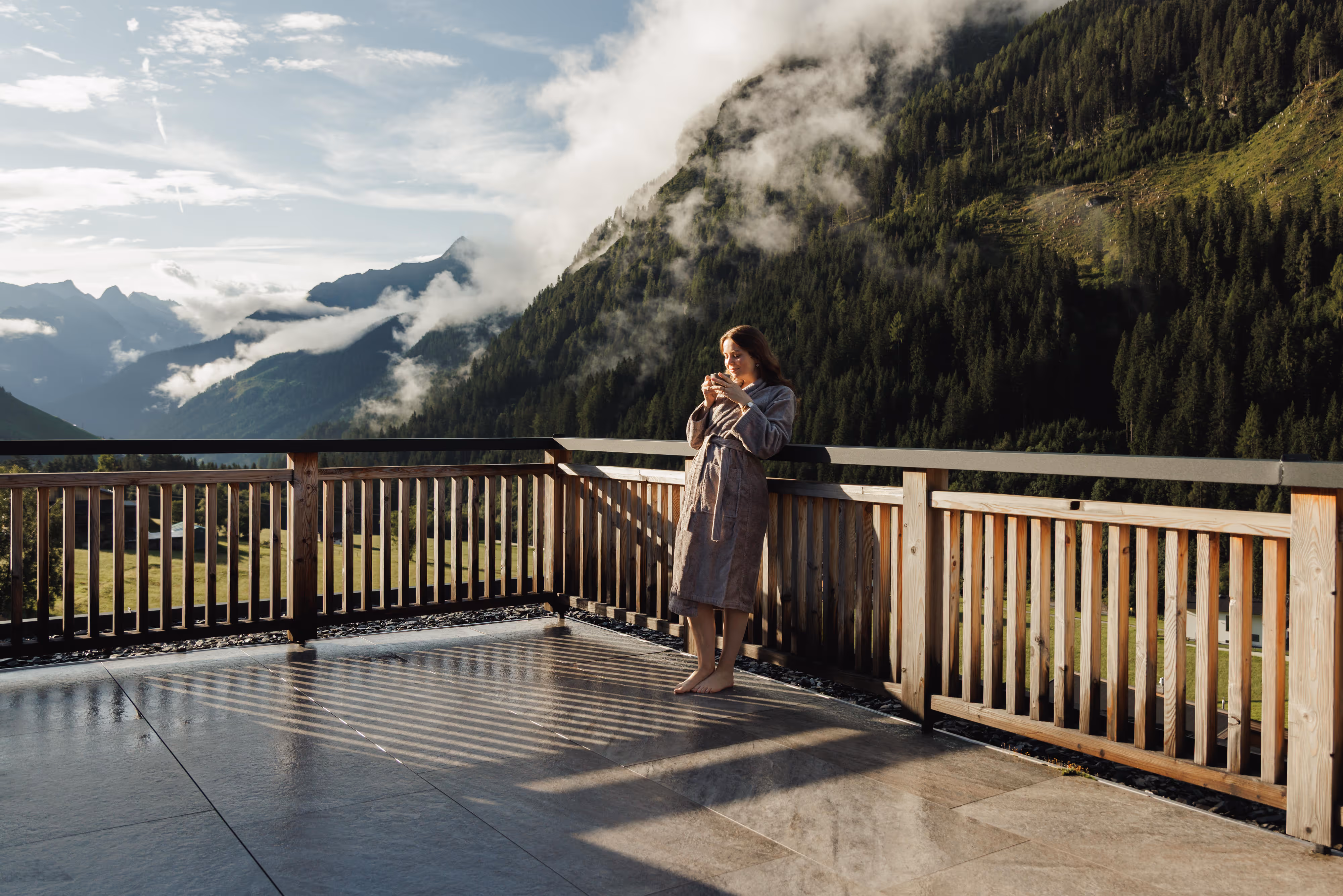 Frau in Bademantel trinkt auf Holzterrasse mit Bergblick und Wolken in der Morgensonne.
