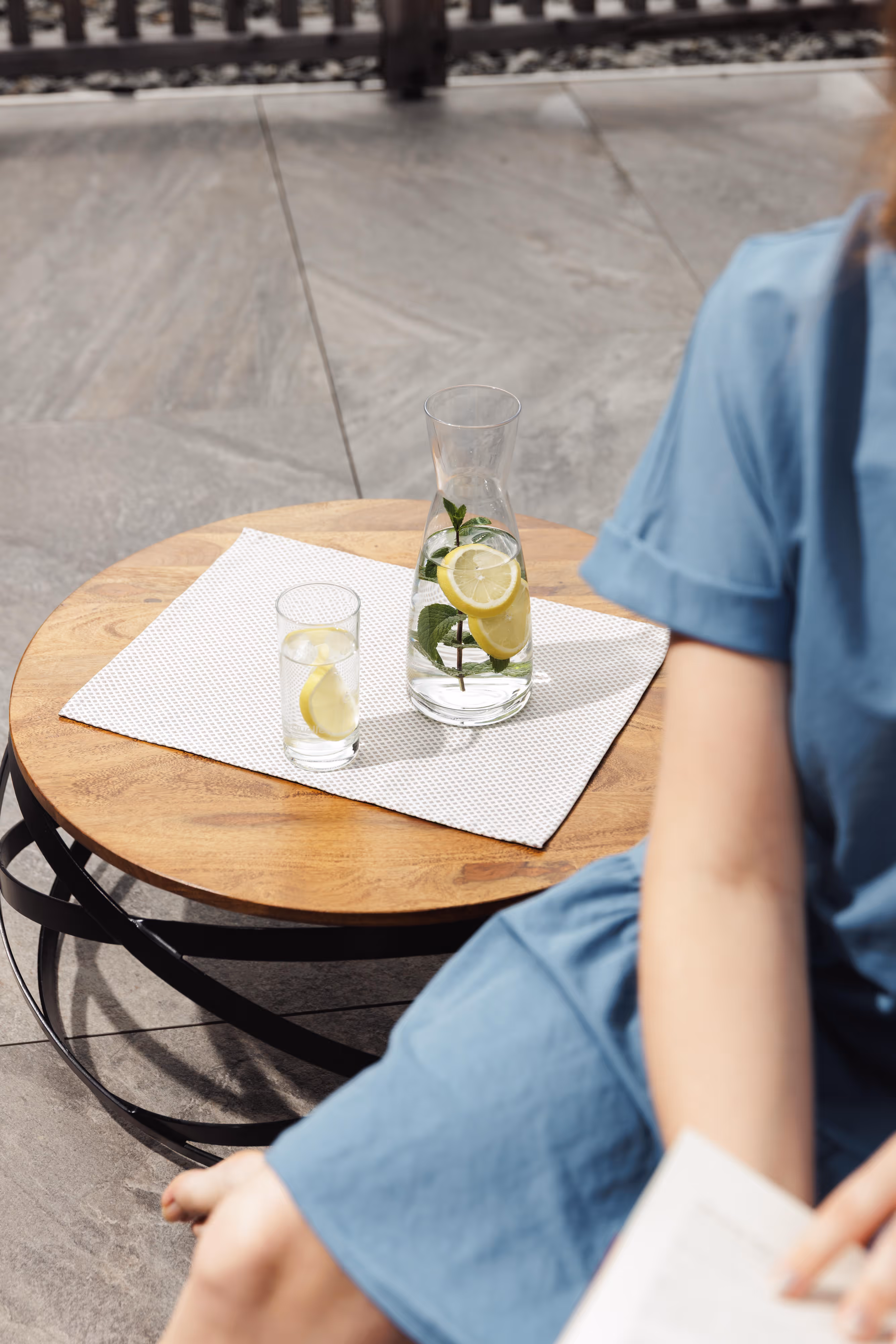 Karaffe und Glas mit Zitronenwasser und Minze auf einem Holztisch neben einer Person im blauen Kleid.