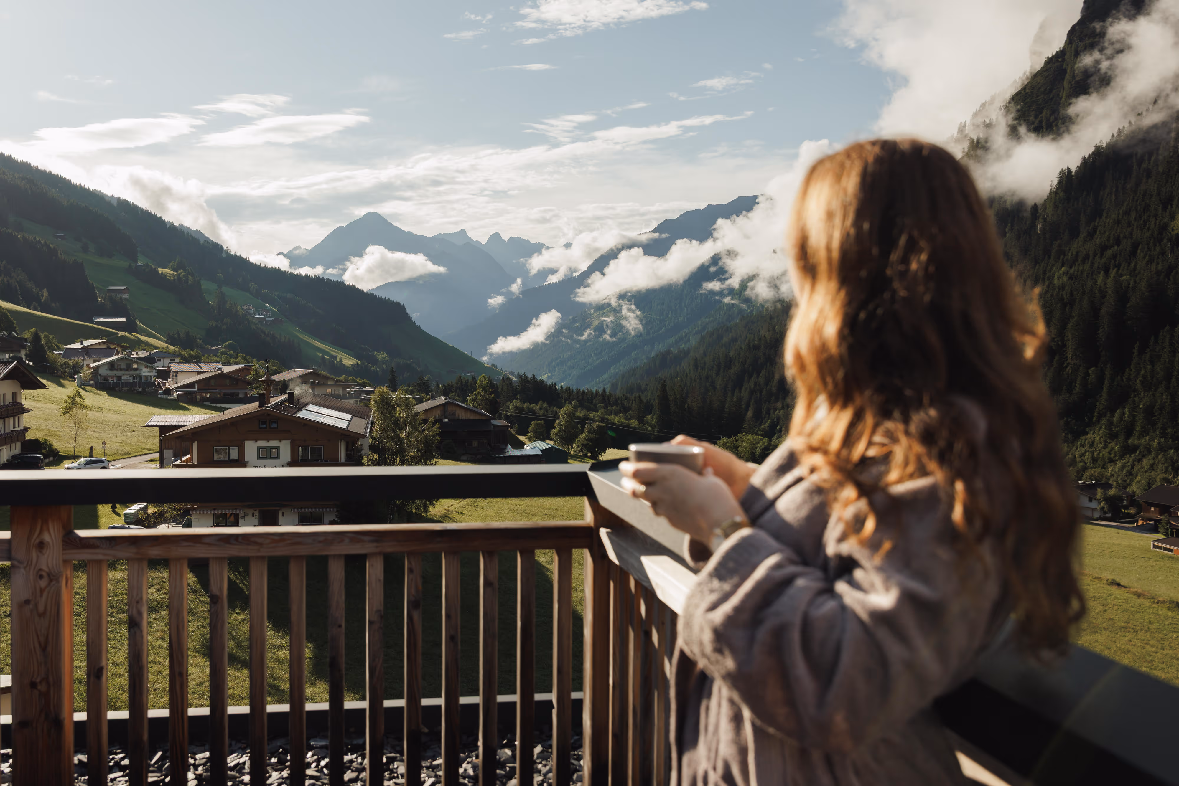 Frau in Morgenmantel hält Tasse auf Balkon mit Alpenblick und Nebel zwischen Bergen.