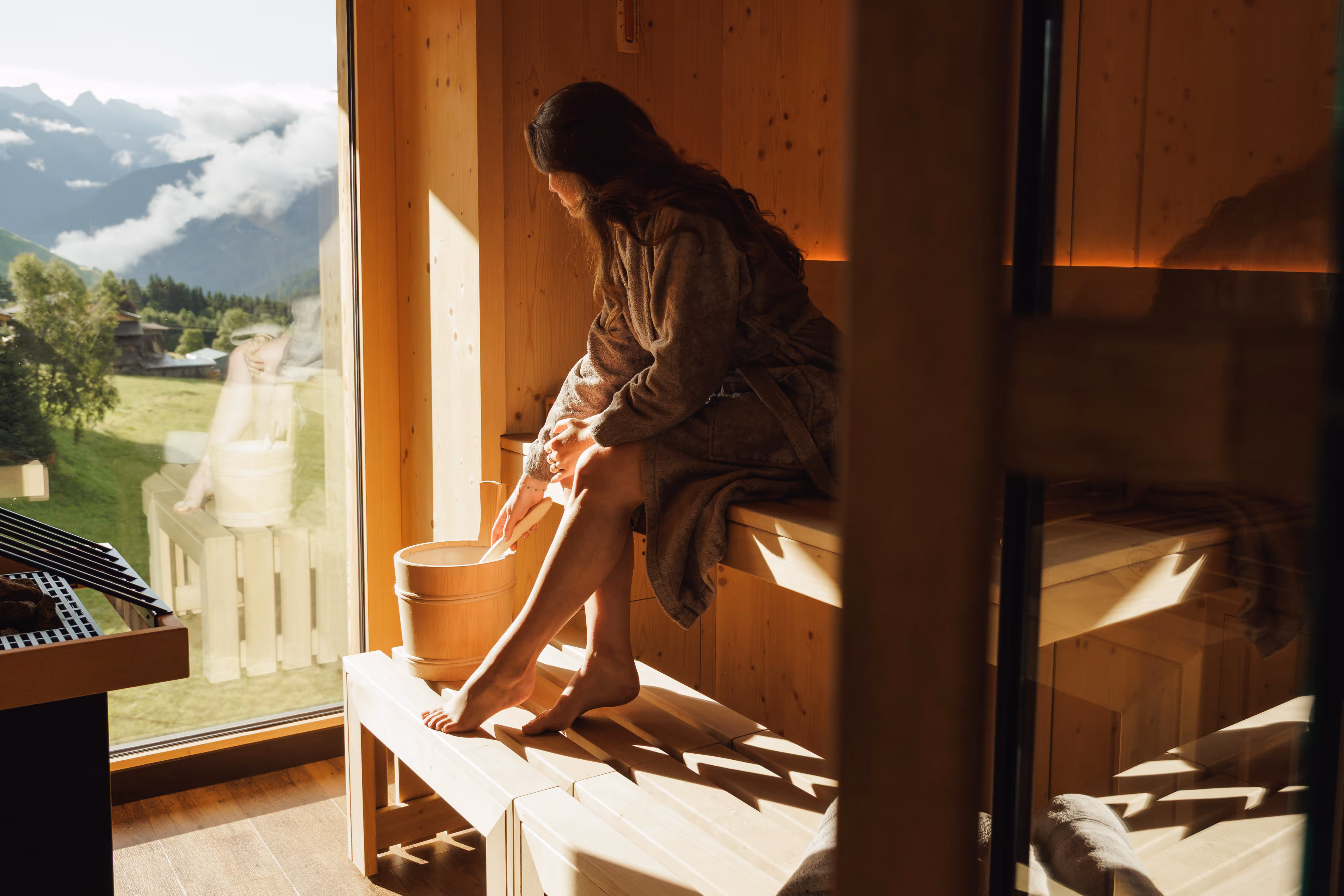 Frau in Bademantel sitzt in Sauna und hält Kelle über einen Holzeimer vor Bergfenster.