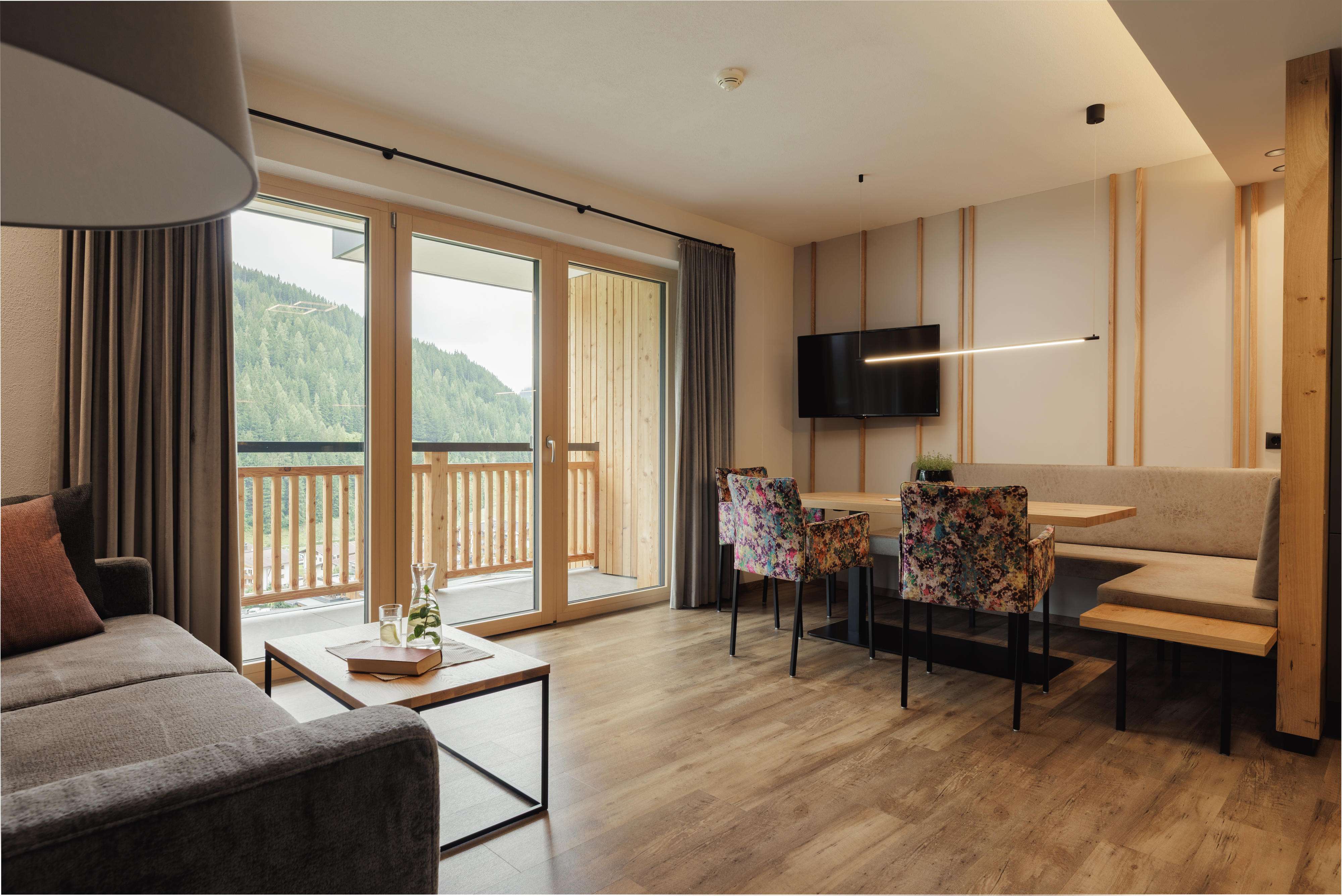 Modernes Wohnzimmer mit Couch, bunten Stühlen, Esstisch und Balkon mit Blick auf bewaldeten Berg.