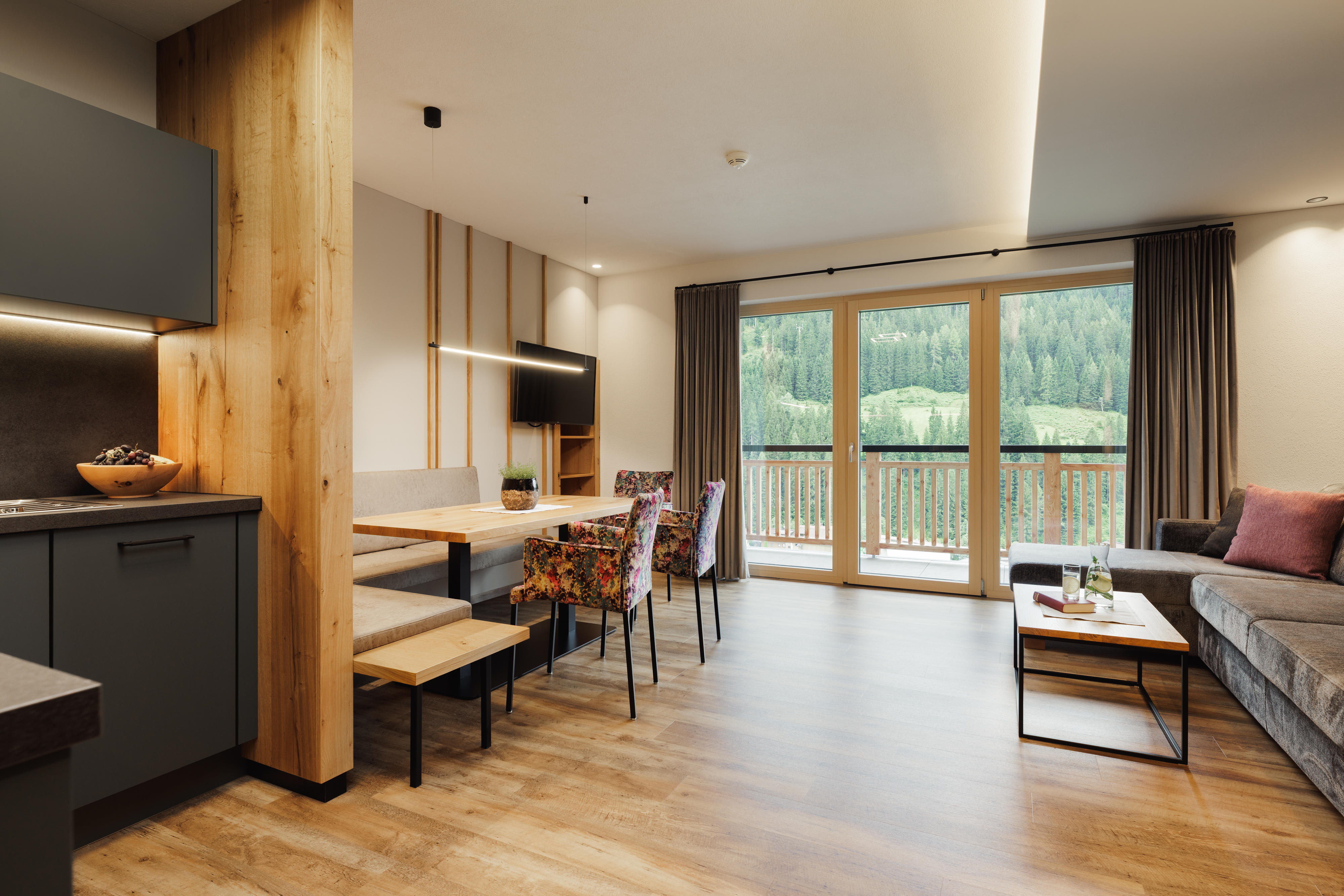 Moderne Wohnküche mit Holzboden, Essbereich, Sofa und großem Fenster mit Blick auf Wald und Berge.