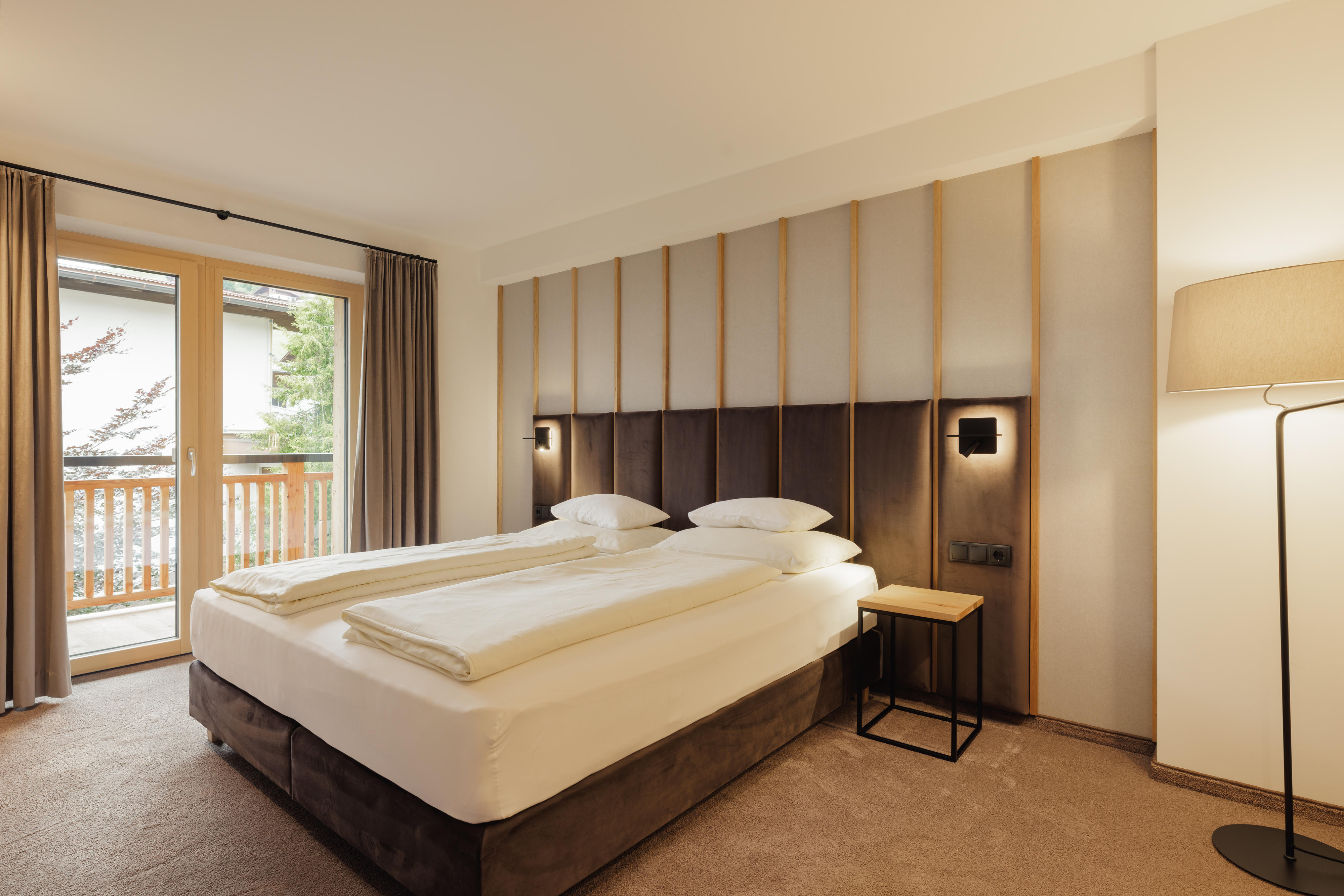 Hell eingerichtetes Schlafzimmer mit Doppelbett, großem Fenster und Balkon im Hintergrund.
