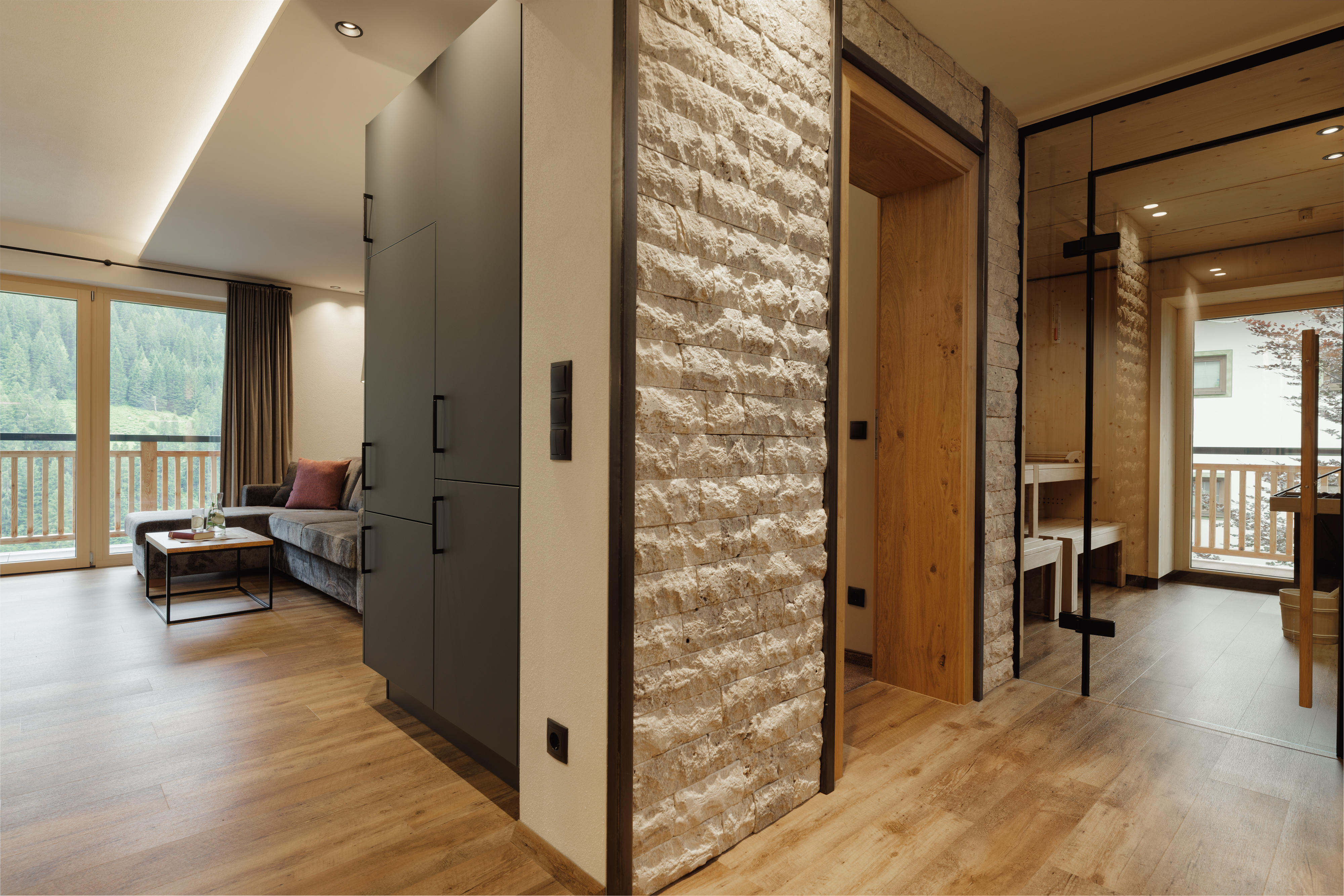 Modernes Wohnzimmer mit Sofa, Steintapete, Holzboden und Sauna hinter Glasschiebetür.