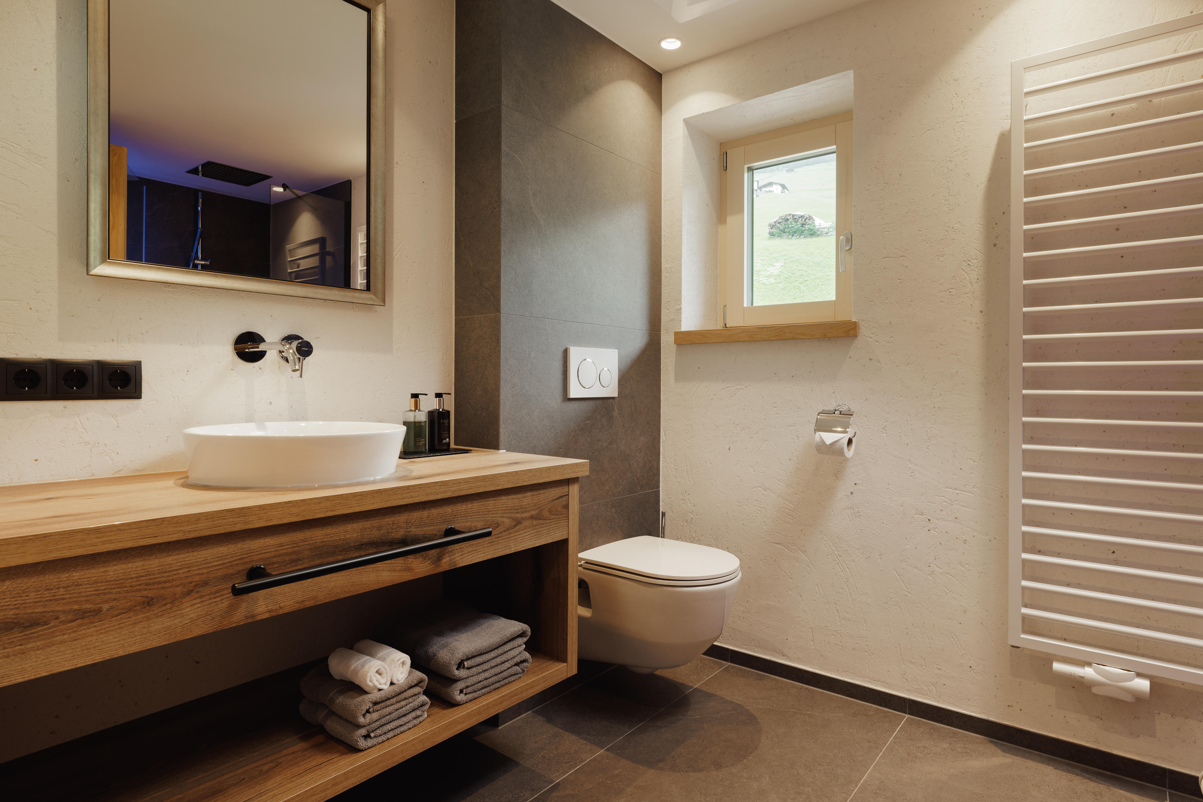 Modern bathroom with wooden vanity, round sink, wall-mounted toilet, small window, and towel radiator.