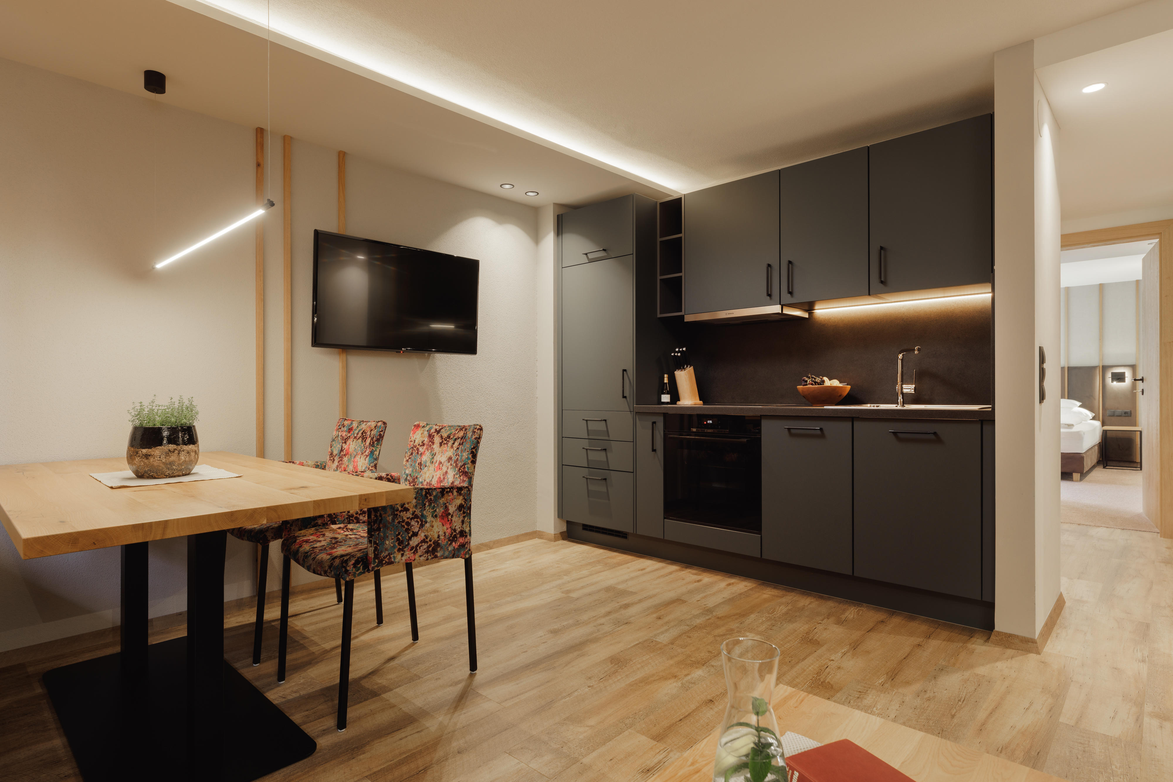 Moderne Küche mit schwarzen Schränken, Holztisch, bunten Stühlen und Wandfernseher.