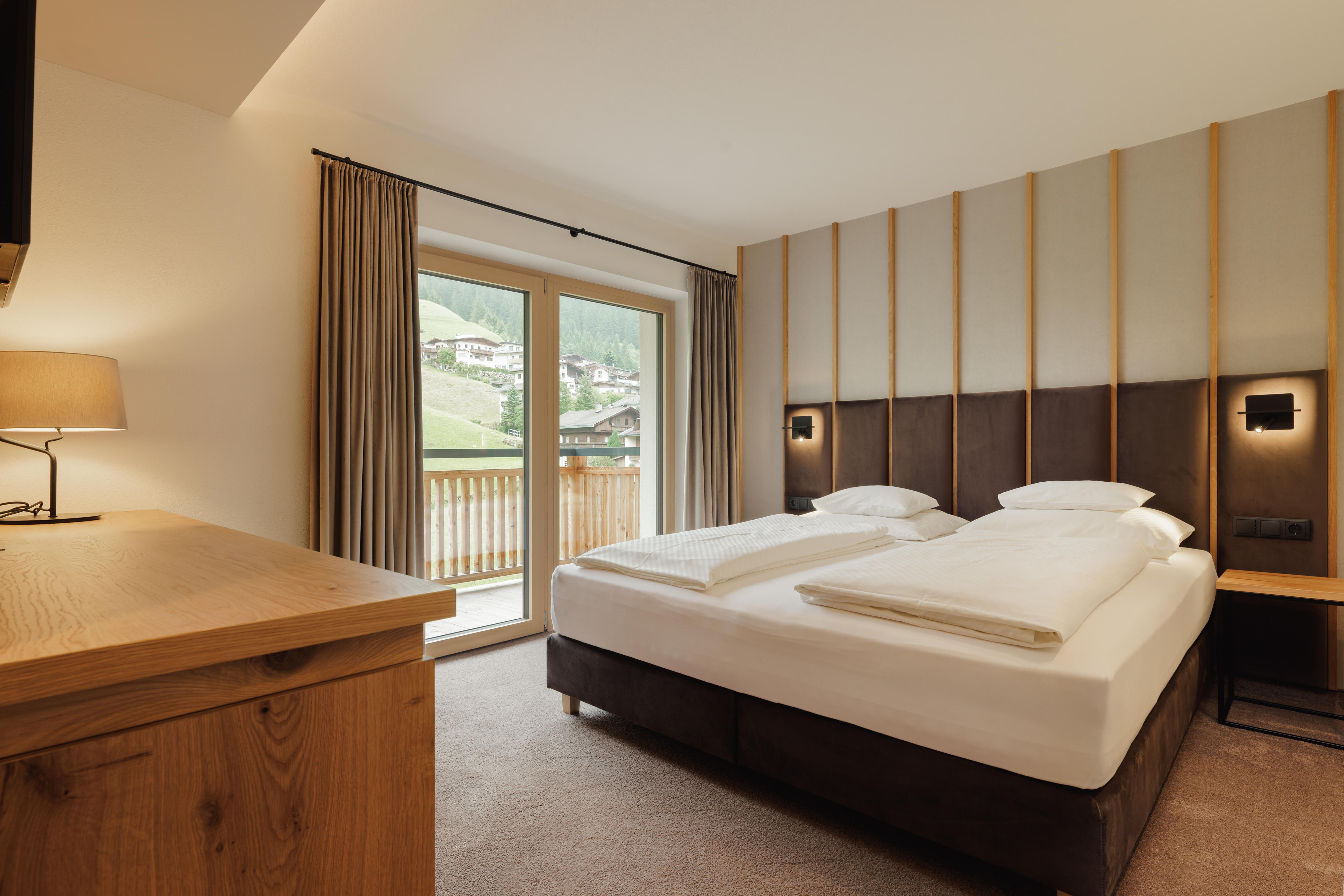 Modern hotel room with double bed, wooden furniture, balcony door, and mountain view outside.
