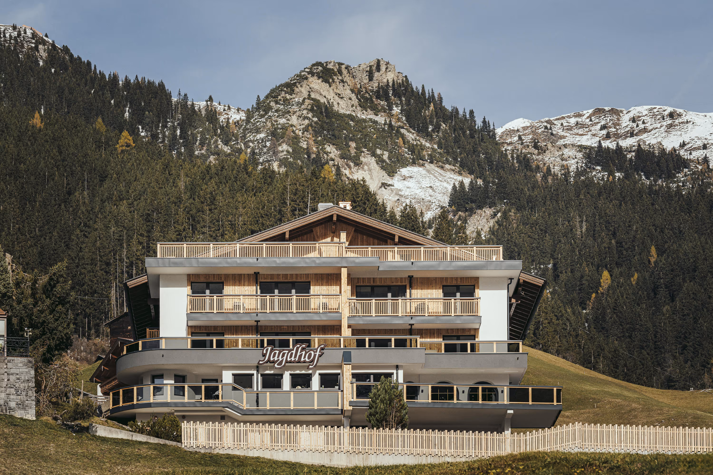 Modern Alpine building Jagdhof with balconies and wood accents, forest and snow-covered mountain behind.
