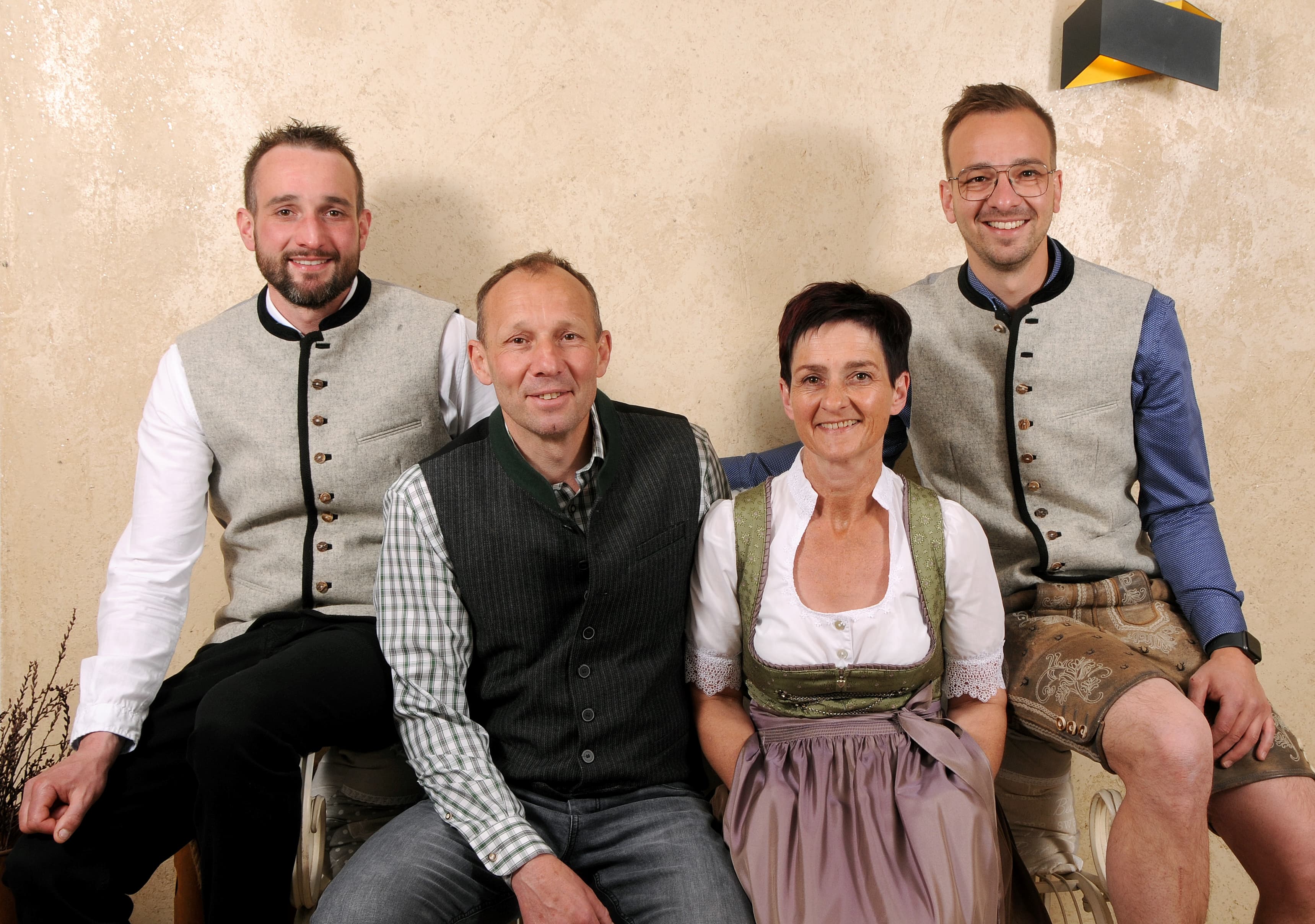 Familie in traditioneller Tracht posiert stehend und sitzend vor beigefarbener Wand, lächelnd.