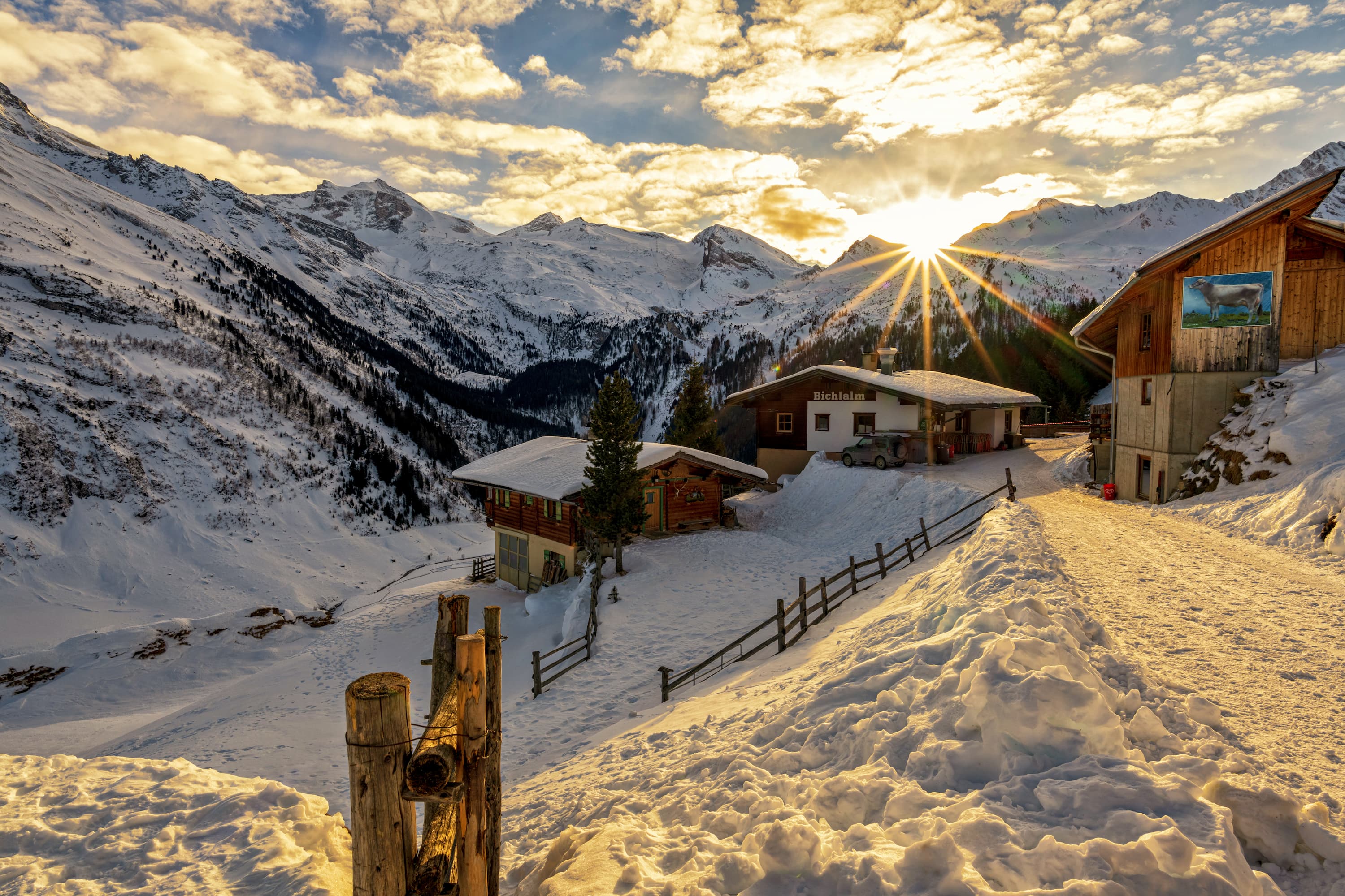 Sonnenaufgang über schneebedeckten Alpenhütten und Bergen mit einem Zaun im Vordergrund.