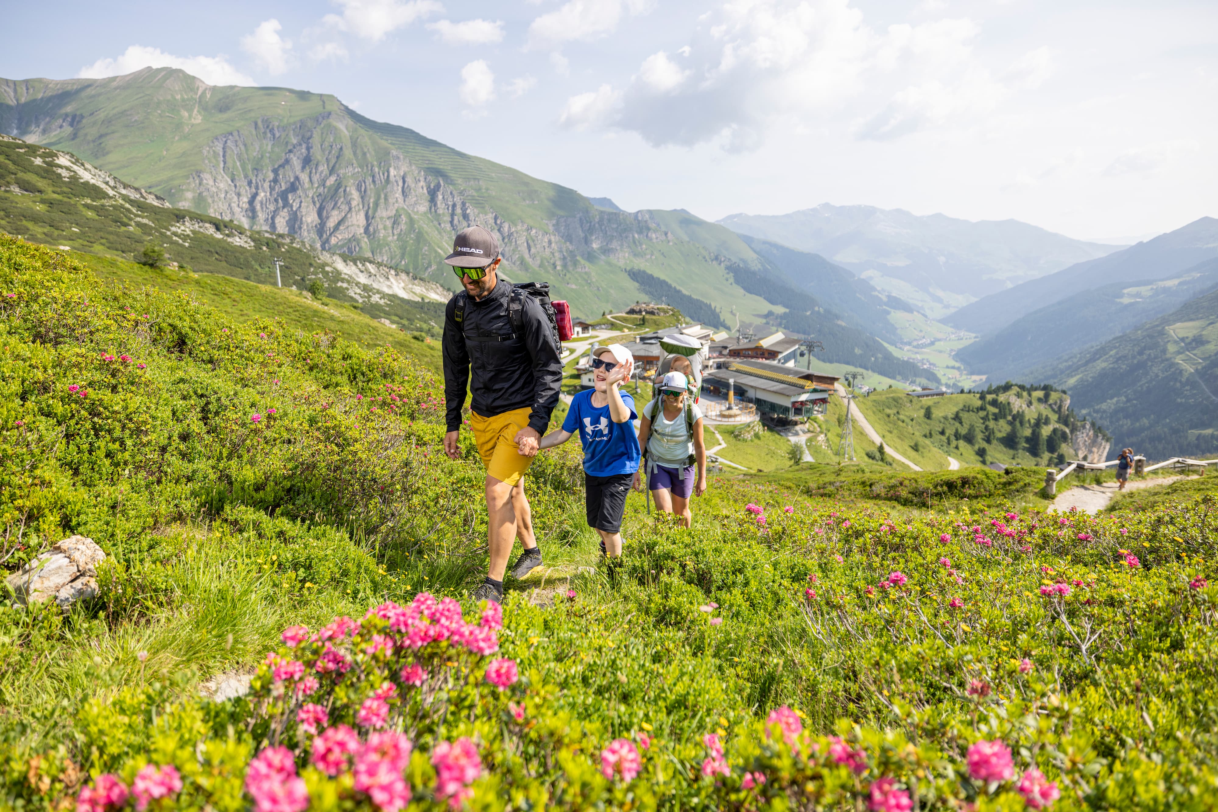 Familie wandert in den Alpen auf einem blumenreichen Bergpfad mit Bahnanlage im Hintergrund.