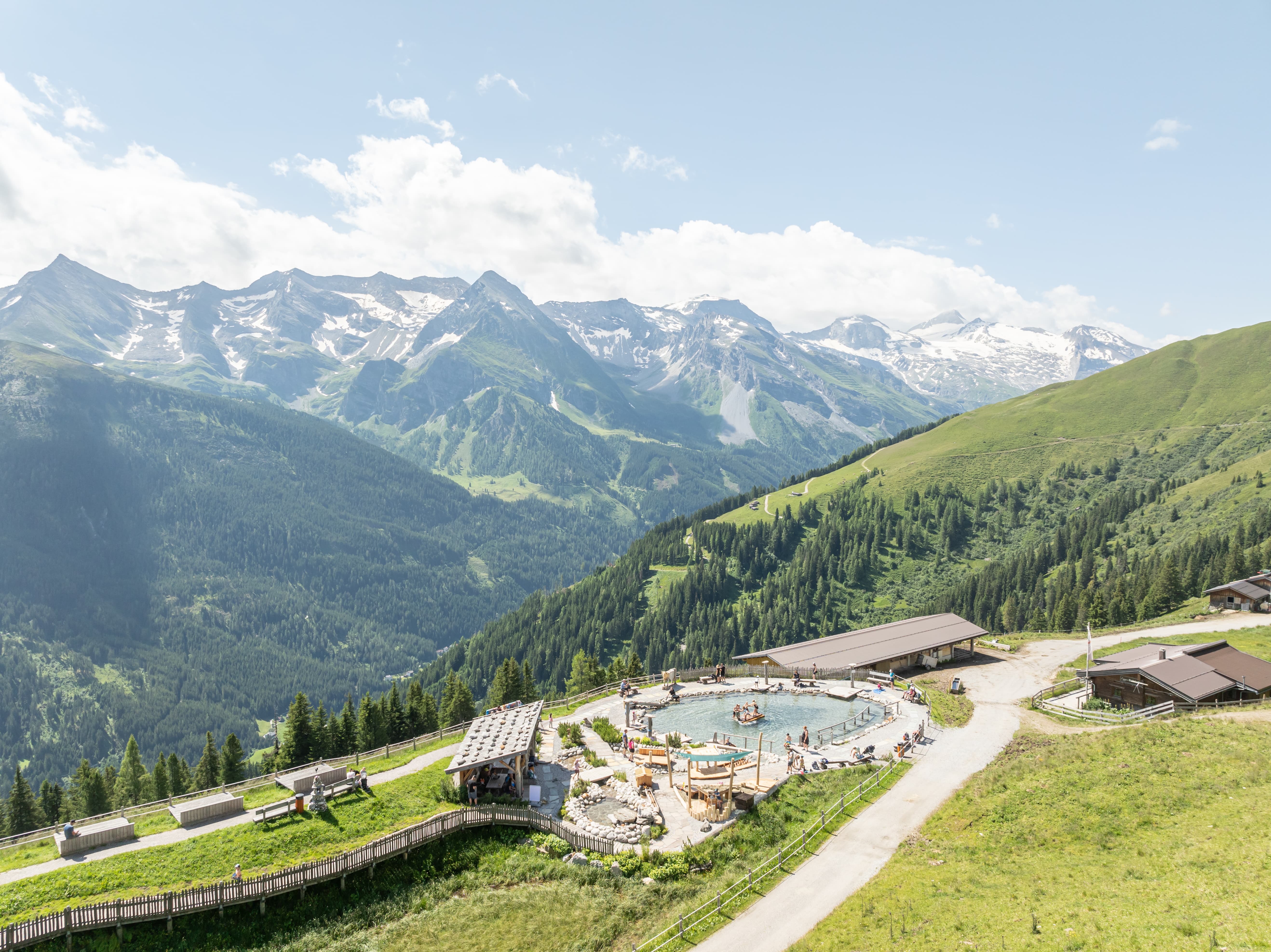 Bergbad mit Menschen, Holzhütten und Bergen mit Schneeflecken in der Ferne an einem sonnigen Tag.