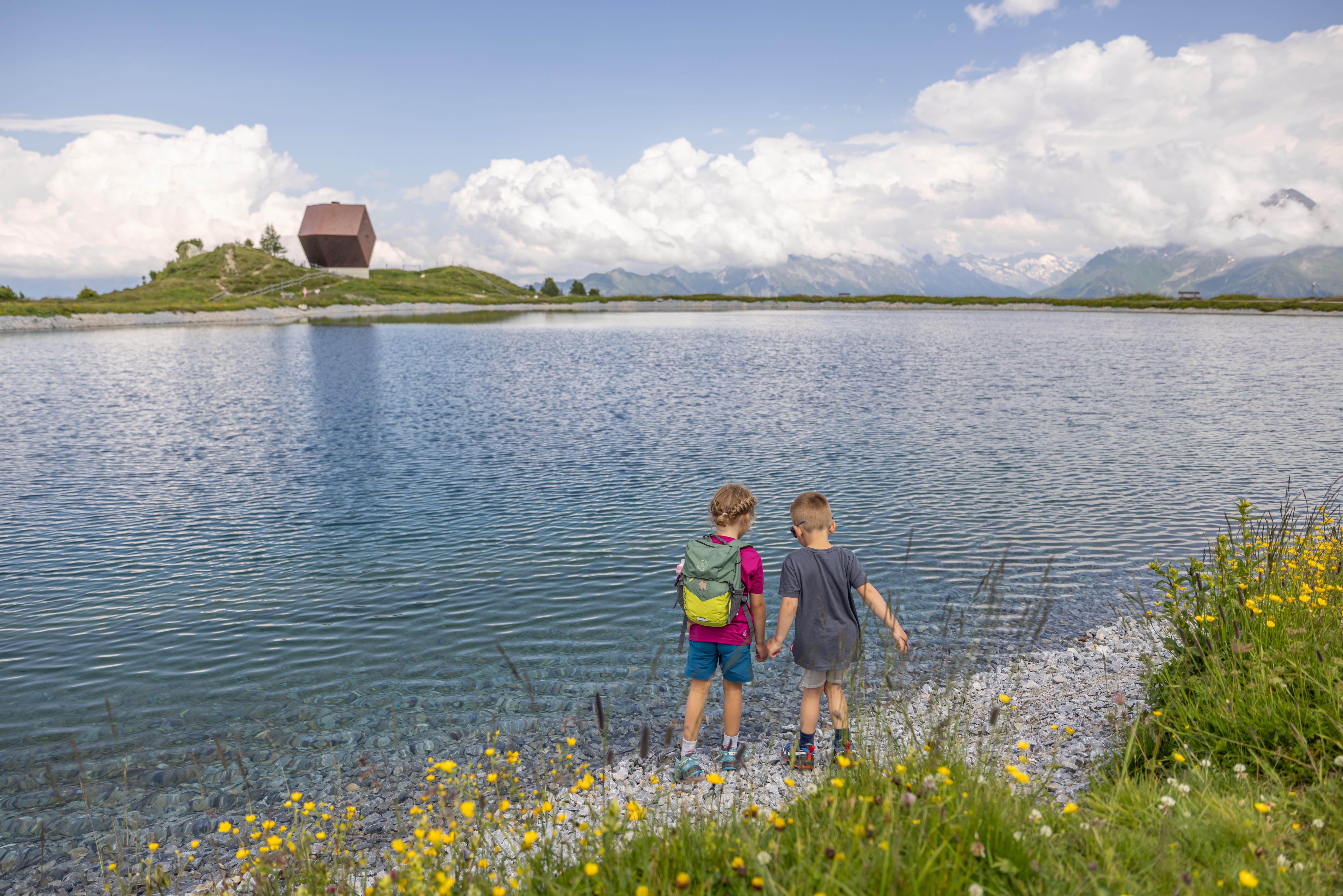 Zwei Kinder halten sich an der Hand am Ufer eines Bergsees mit Blumen und Bergen im Hintergrund.