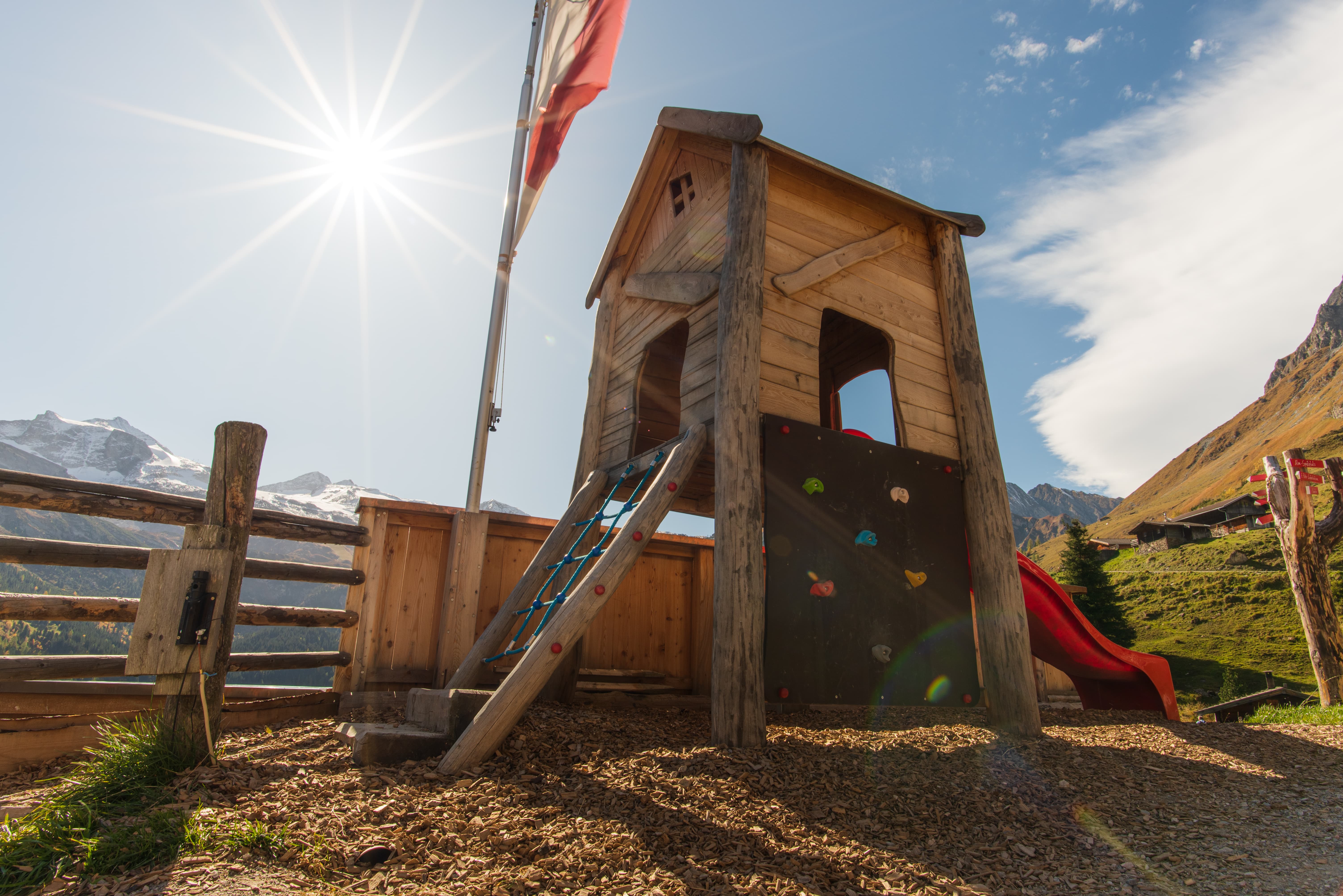 Kinderspielhaus aus Holz mit Kletterwand und Rutsche vor einer Berglandschaft im Sonnenschein.